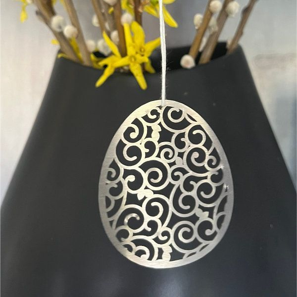 Ein silbernes Filigran-Ei hängt vor einer dunklen Vase mit gelben Blumen.