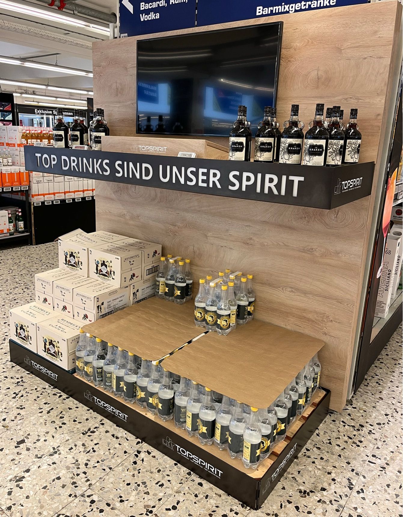 Getränkepräsentation in einem Geschäft. Flaschen und Kartons sind in Regalen angeordnet, dazu der Text „Top drinks sind unser Spirit“.