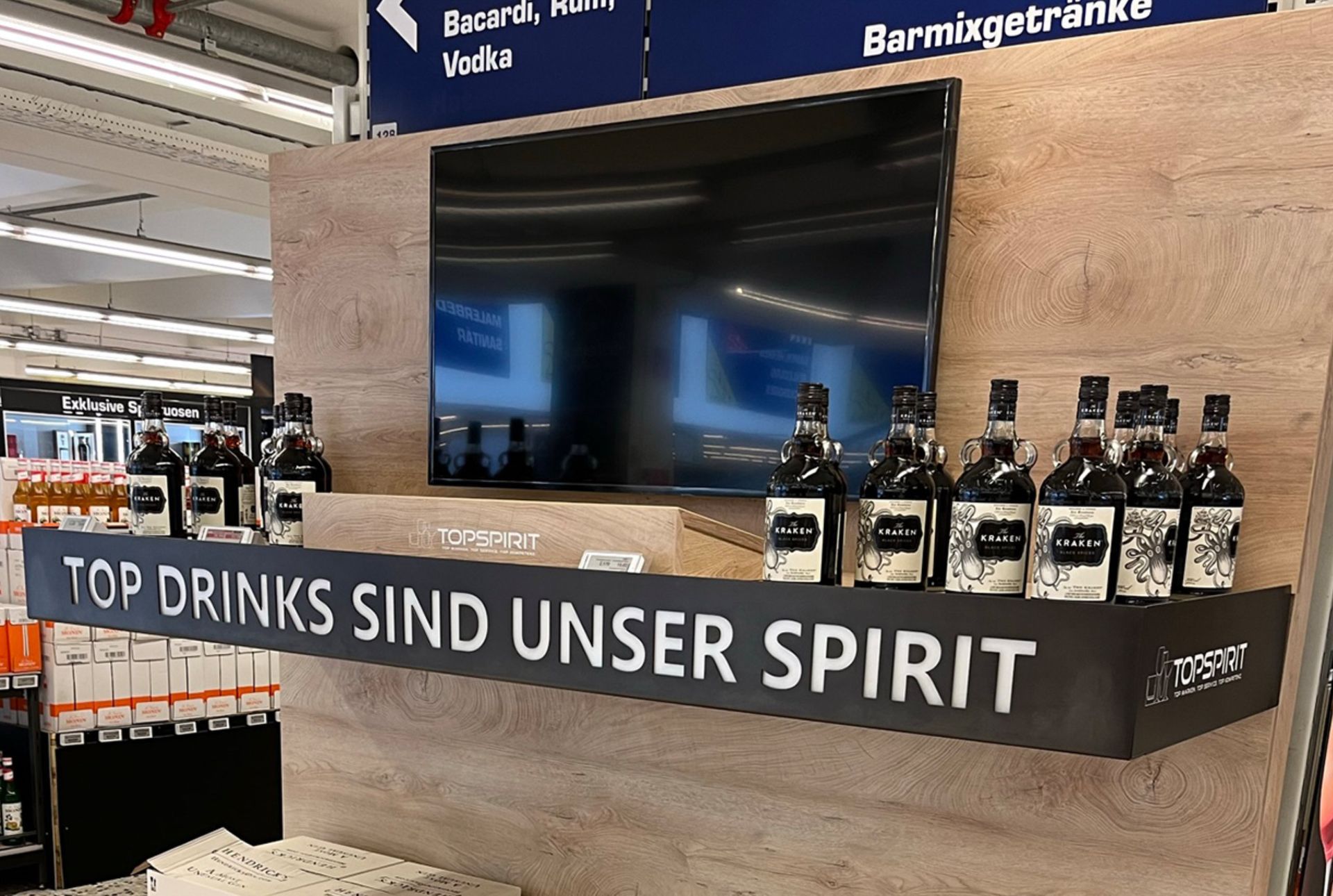 In einem Laden stehen Flaschen mit Spirituosen in einem Regal, darüber hängt ein Fernsehbildschirm. Der Text lautet: „Top Drinks sind unser Spirit.“