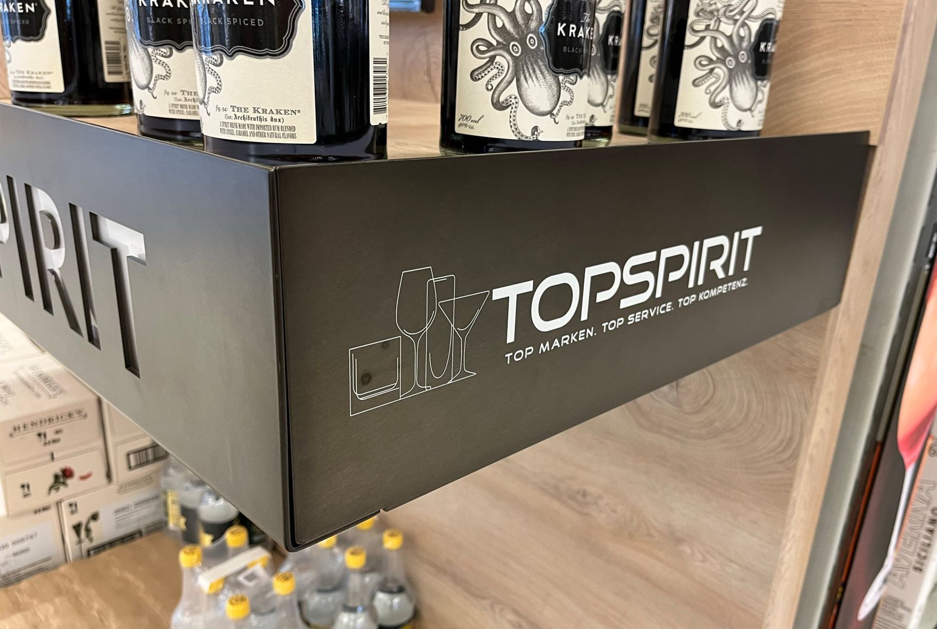 Schwarzes Schild mit dem Logo „TOPSPIRIT“, darüber Spirituosenflaschen in einer Ladenumgebung.