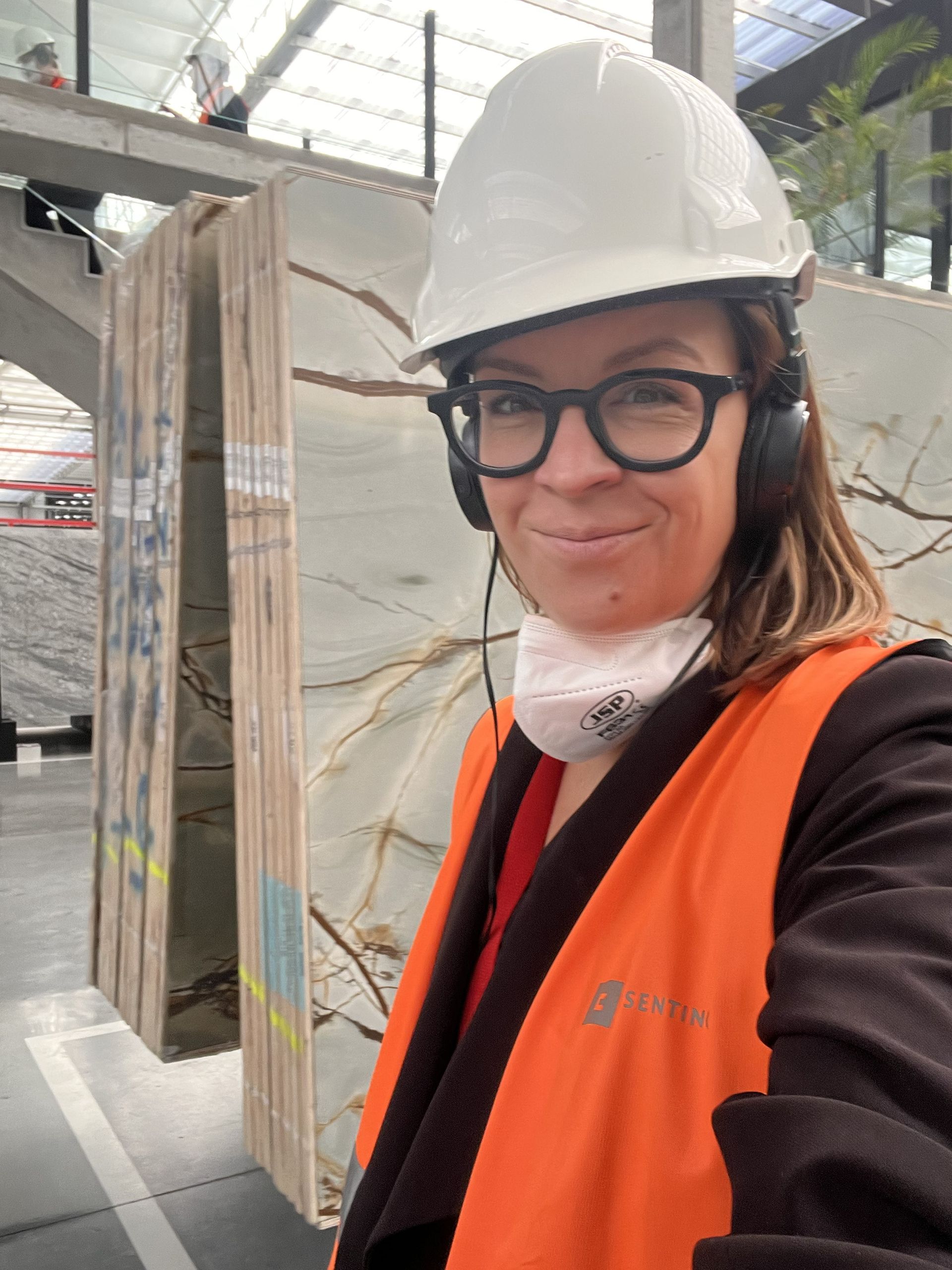 Eine Frau mit Schutzhelm, Sicherheitsweste und Brille lächelt in einer Fabrikhalle; hinter ihr sind große Steinplatten zu sehen.