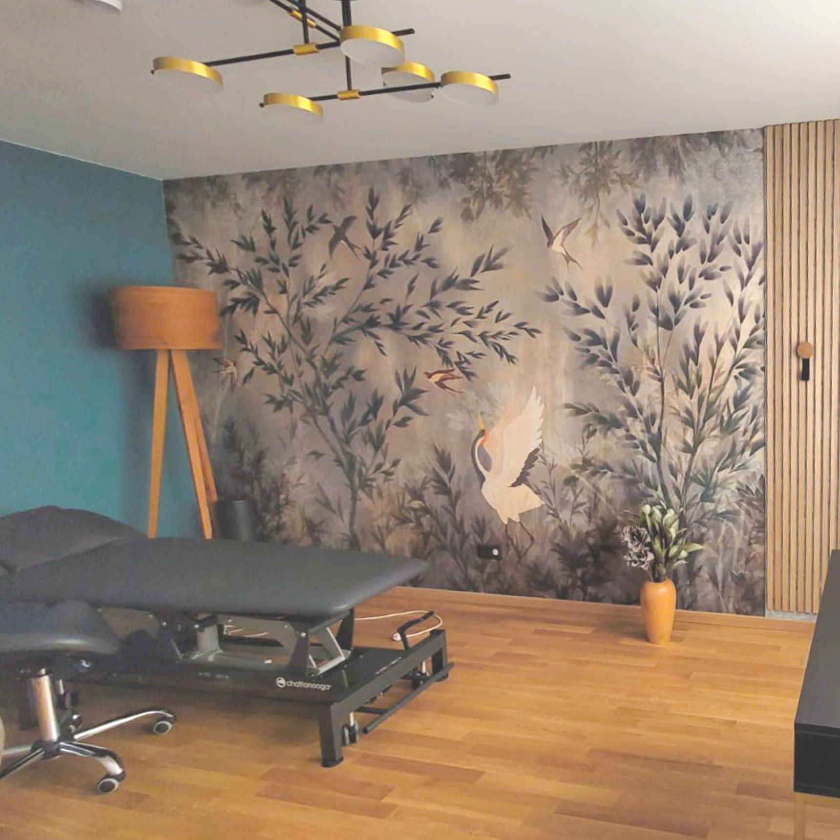 Therapieraum mit Massageliege, Naturwandbild, Holzboden und moderner Beleuchtung.
