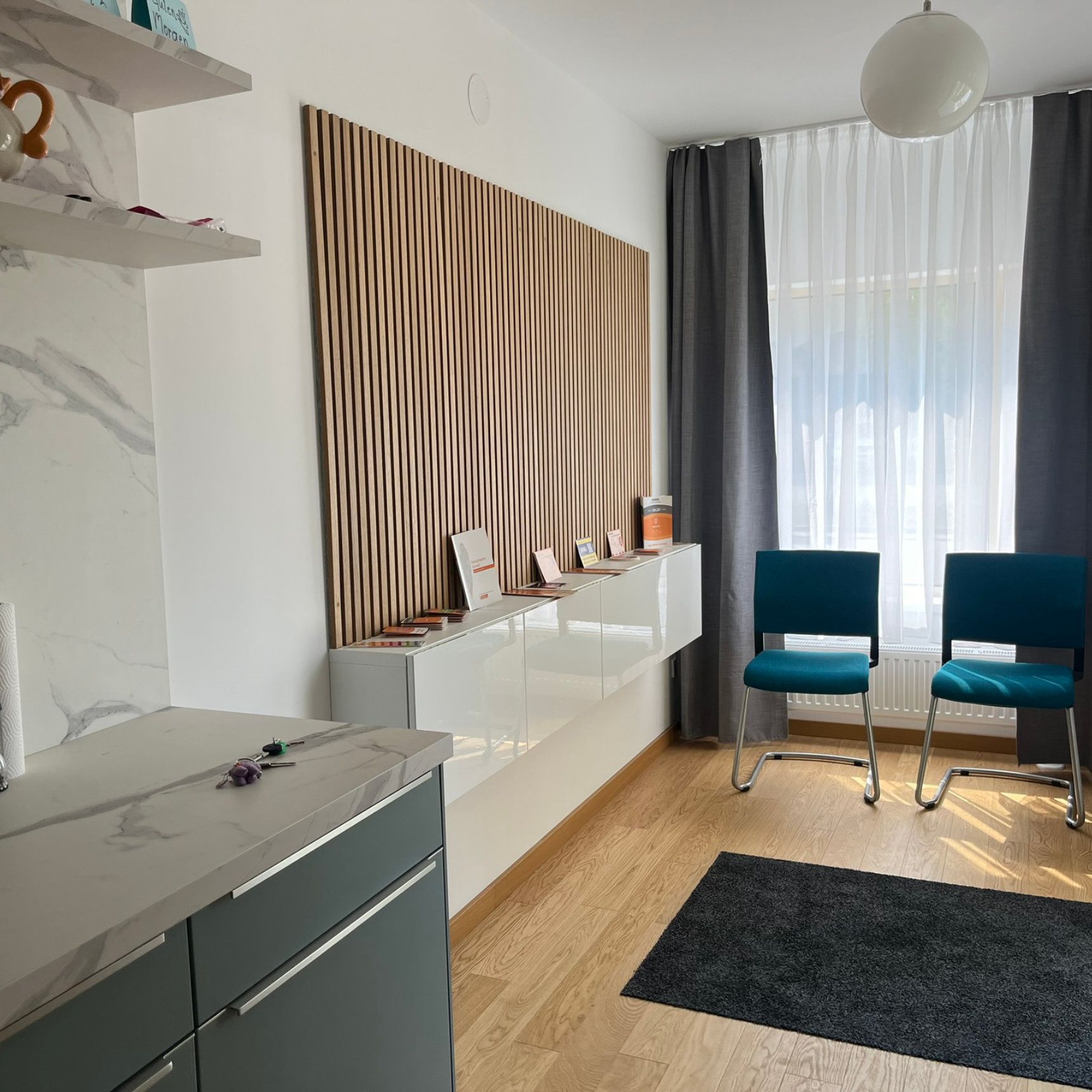 Wartezimmer mit zwei türkisfarbenen Stühlen, hölzerner Wanddekoration und einem Fenster mit grauen Vorhängen.