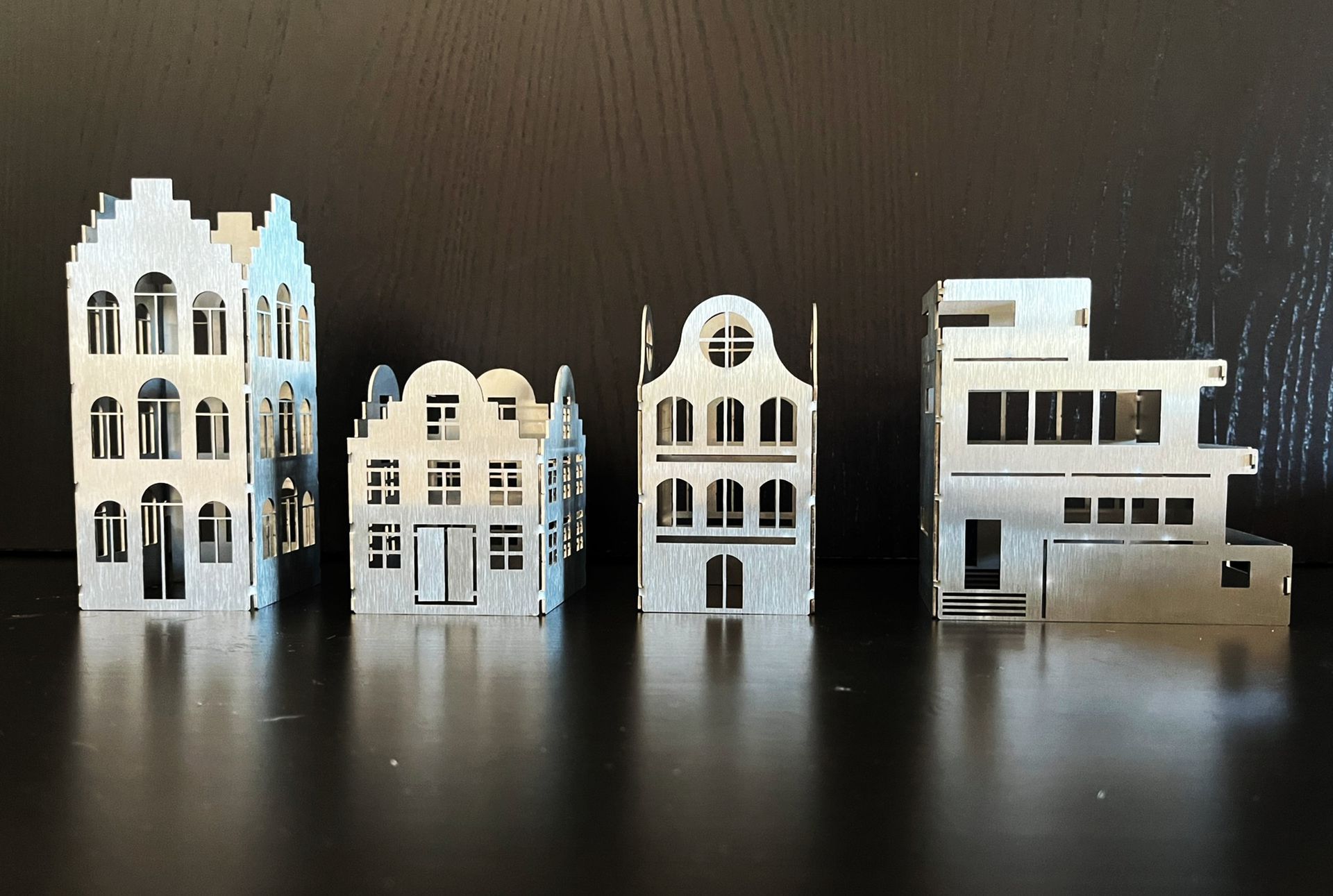 Vier Miniaturhäuser aus silbernem Metall, die auf einer spiegelnden Oberfläche angeordnet sind.