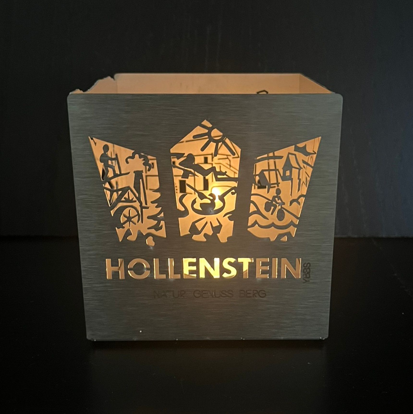 Eine brennende Kerze in einer quadratischen Laterne mit dem Namen „HOLLENSTEIN“ und einer Silhouette auf der Vorderseite.
