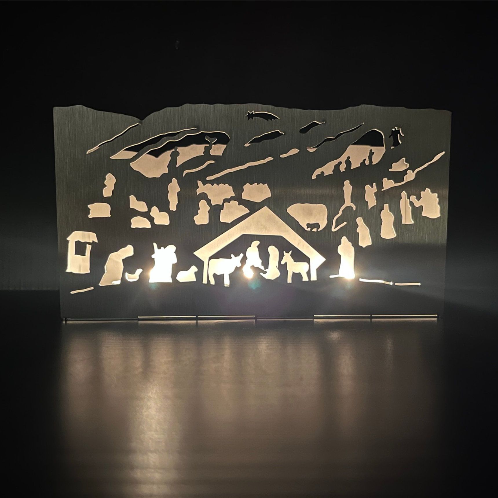 Beleuchtete Silhouette einer Krippe aus Metall mit Tieren, Figuren und Stall; strahlt Licht aus.