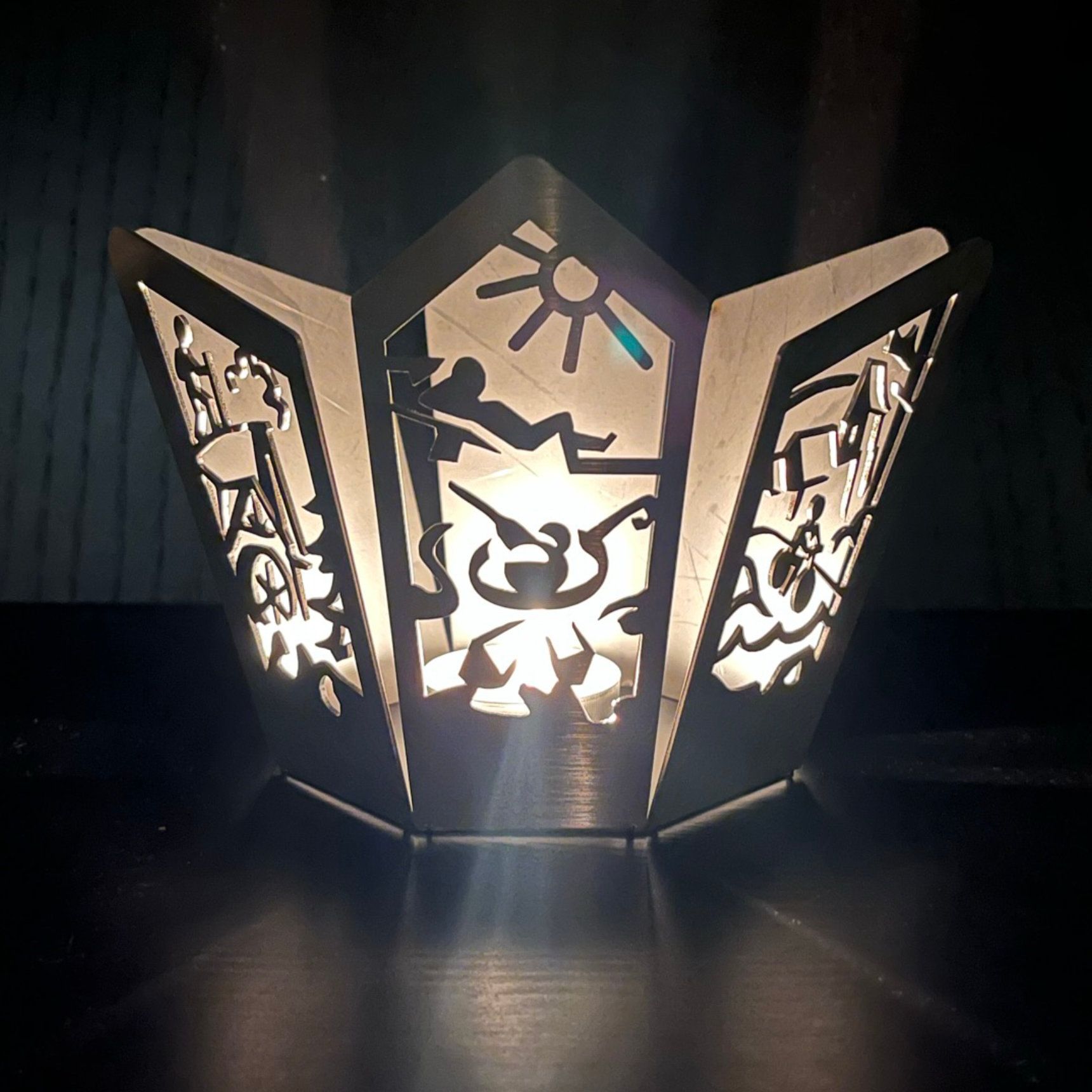 Metalllaterne mit ausgeschnittenen Motiven, von innen beleuchtet. Zeigt eine Strandszene: Sonne, Figuren und Vegetation.