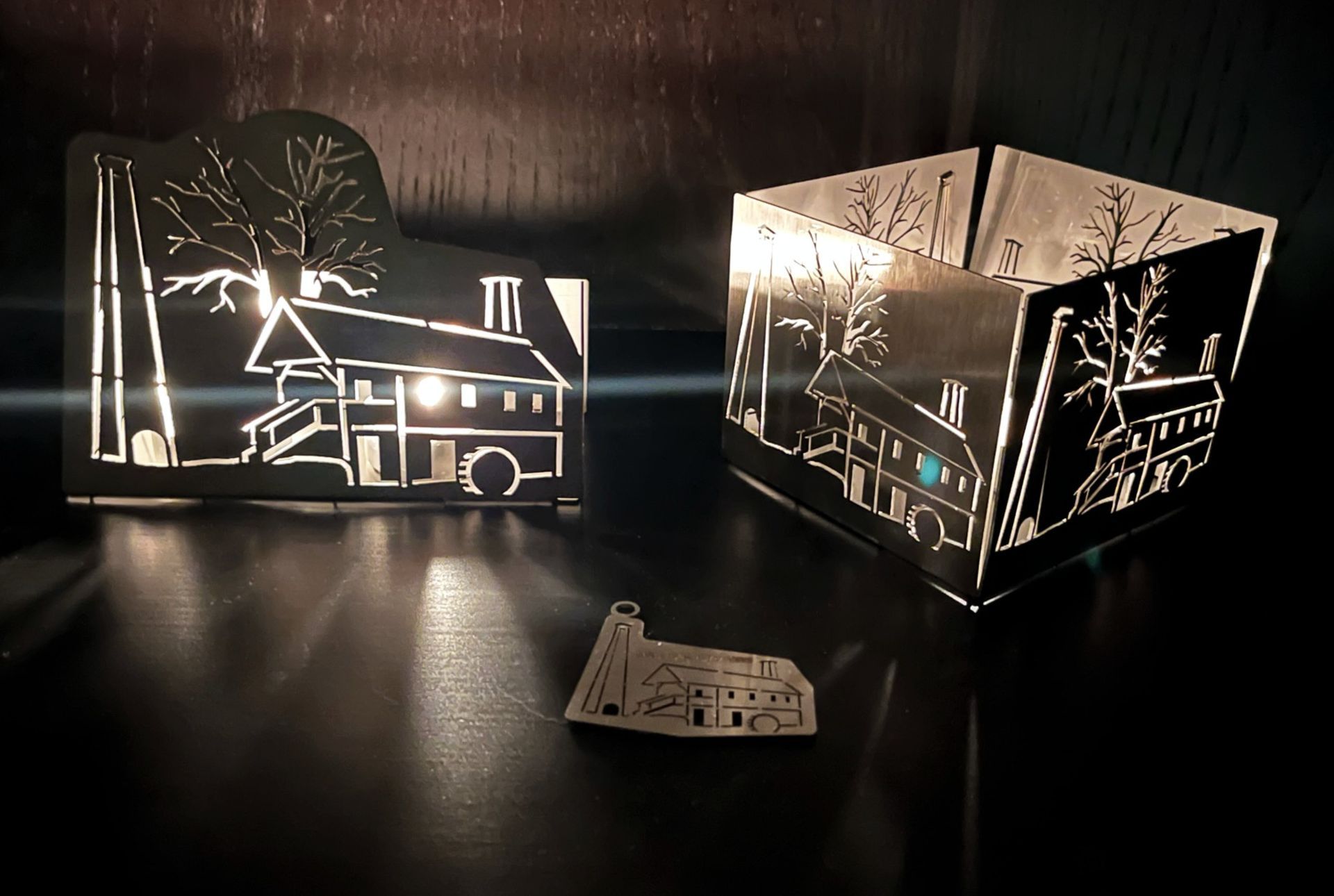 Metallene, hausförmige Kerzenhalter mit ausgeschnittenen Mustern, die vor einem dunklen Hintergrund beleuchtet sind.