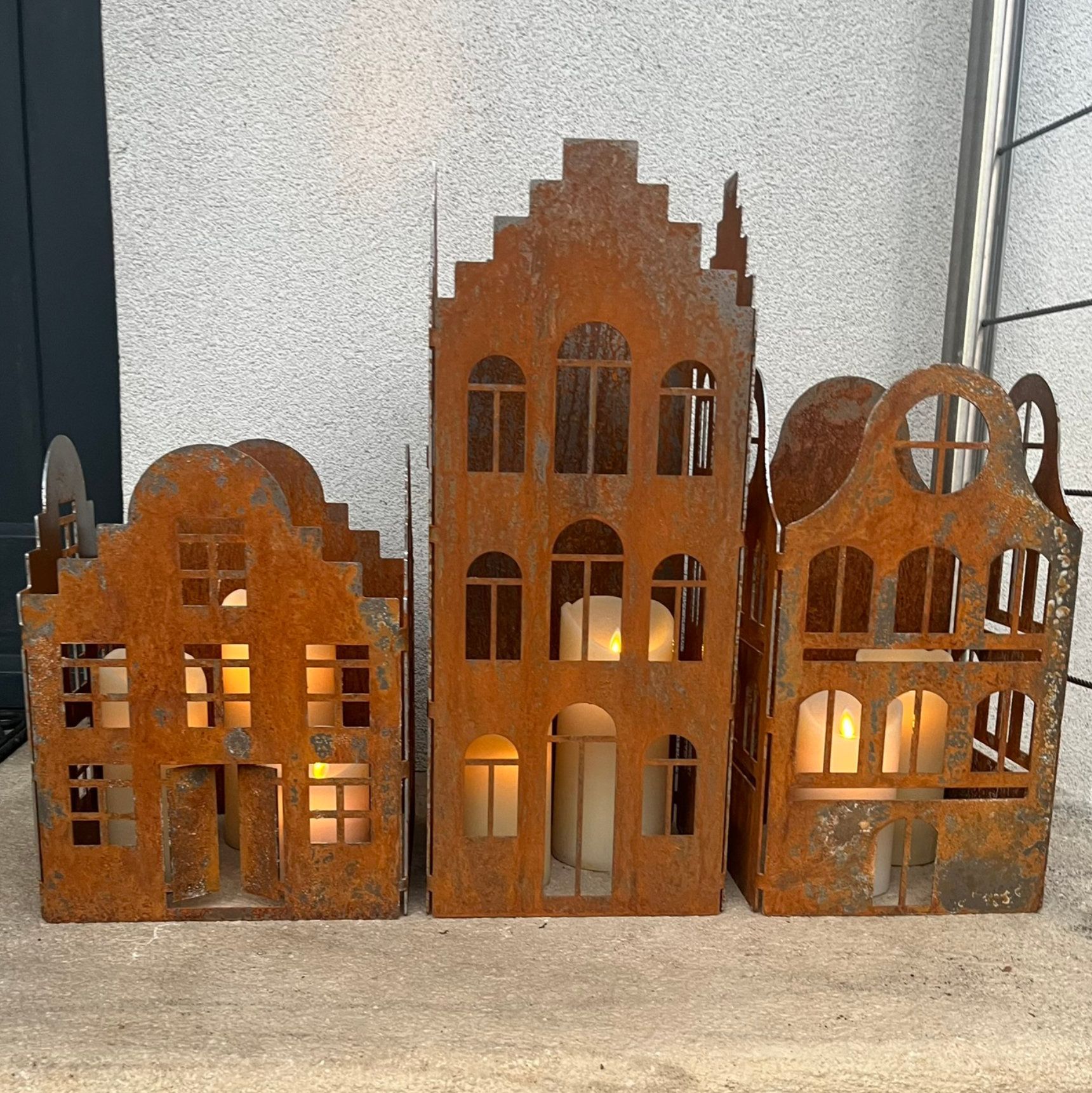 Drei rostige, hausförmige Kerzenhalter aus Metall mit brennenden Kerzen darin vor einem Betonhintergrund.