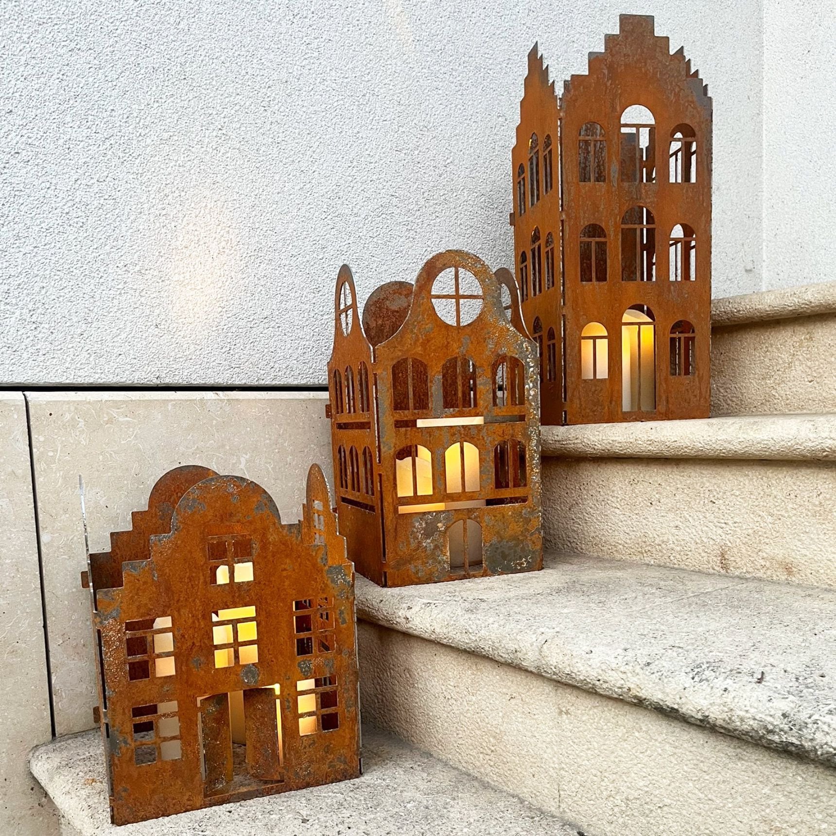 Drei rostige, hausförmige Kerzenhalter aus Metall auf Steinstufen. Brennende Kerzen leuchten darin.