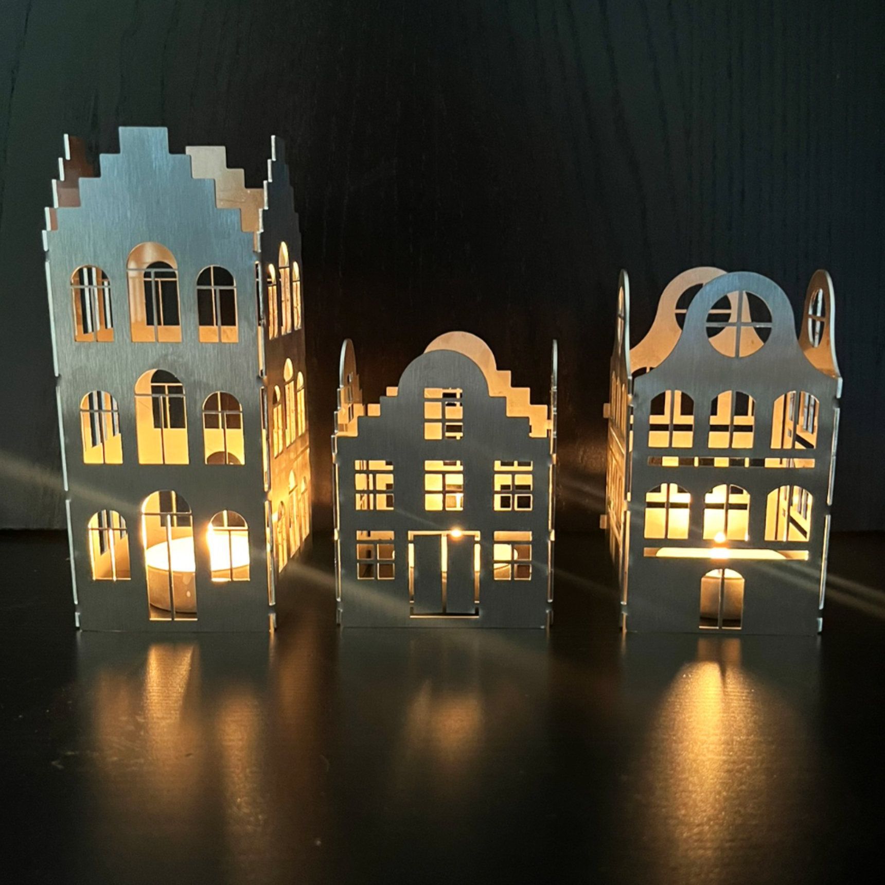 Drei lasergeschnittene, hausförmige Kerzenhalter aus Holz mit brennenden Kerzen im Inneren, die warmes Licht spenden.