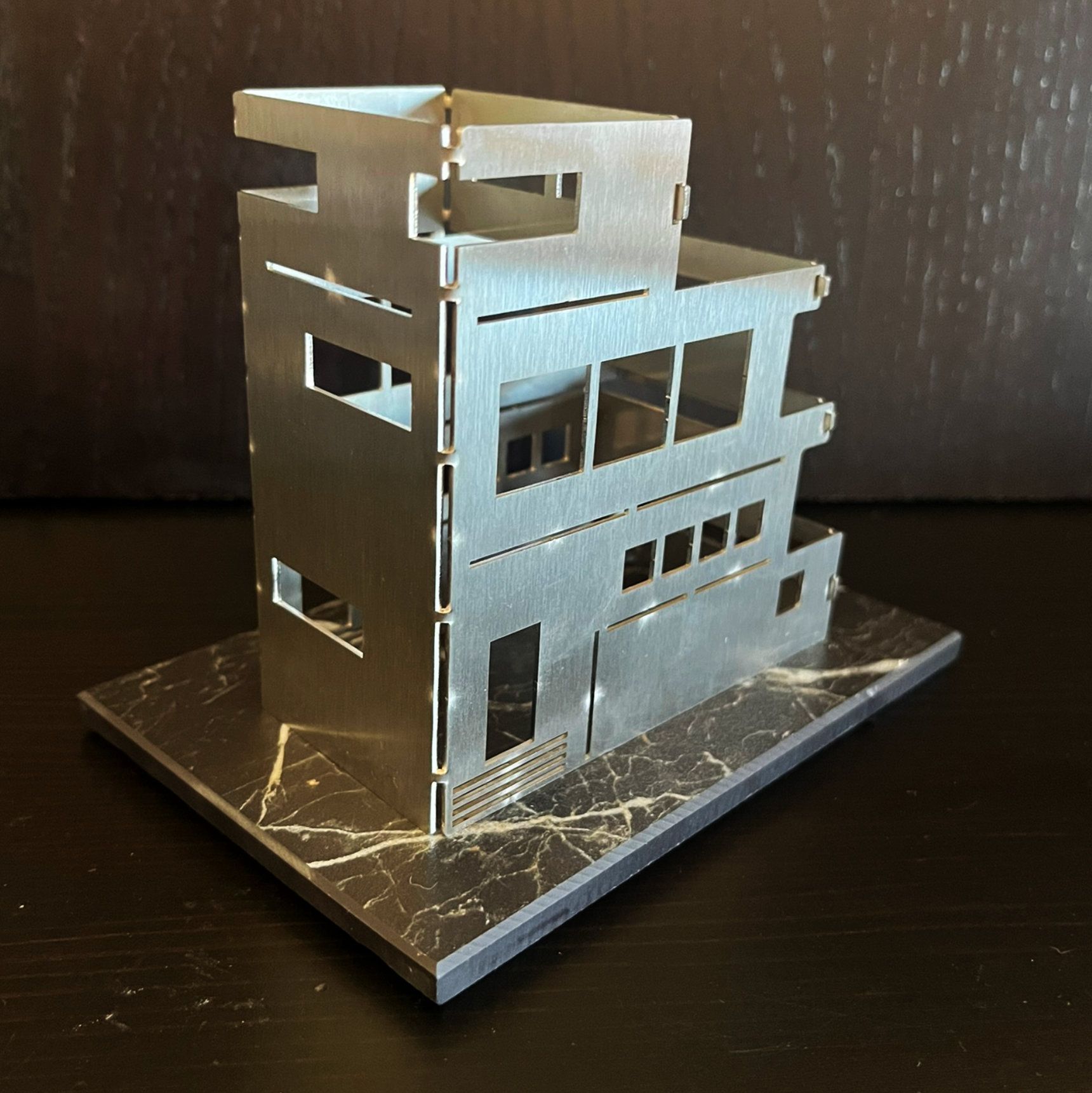 Architekturmodell eines mehrstöckigen Gebäudes aus silbernem Metall mit ausgeschnittenen Fenstern auf strukturiertem Sockel.
