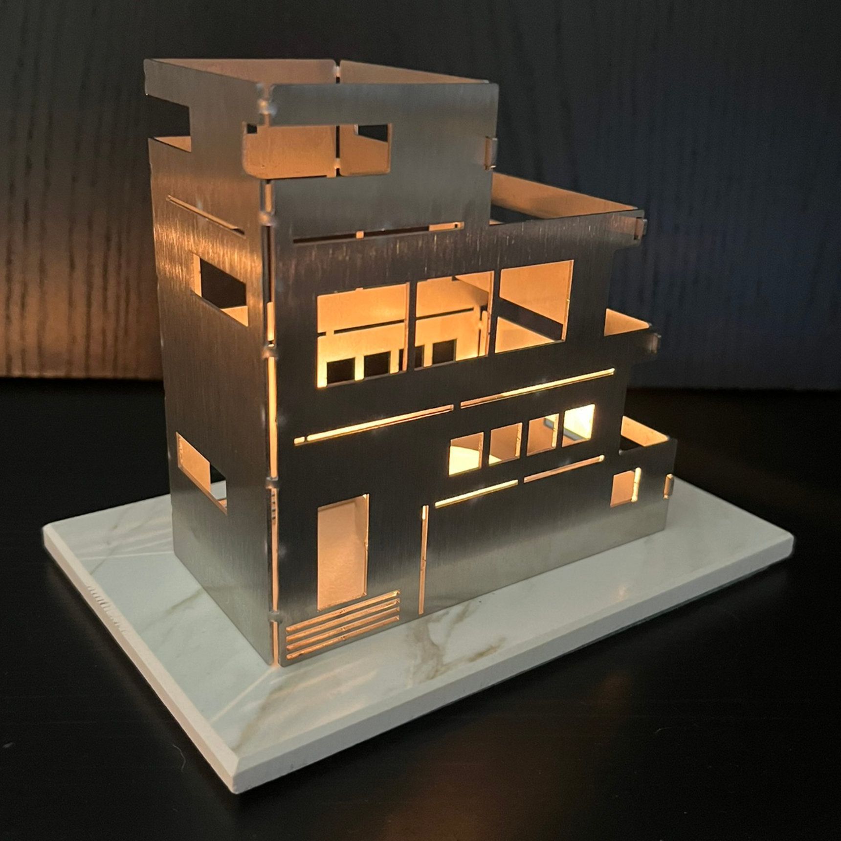 Metallmodell eines modernen Gebäudes mit beleuchteten Innenfenstern, auf einem hellen Holzsockel.