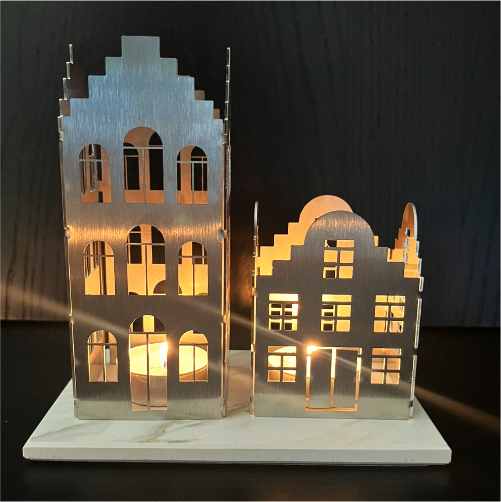 Zwei metallene, hausförmige Kerzenhalter mit brennenden Kerzen, die auf einem weißen Steinsockel stehen.