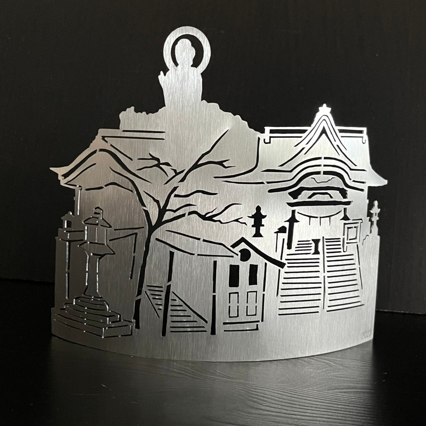 Metallausschnitt einer japanischen Tempelszene mit Gebäuden, Bäumen und einer Buddha-Figur an der Spitze.