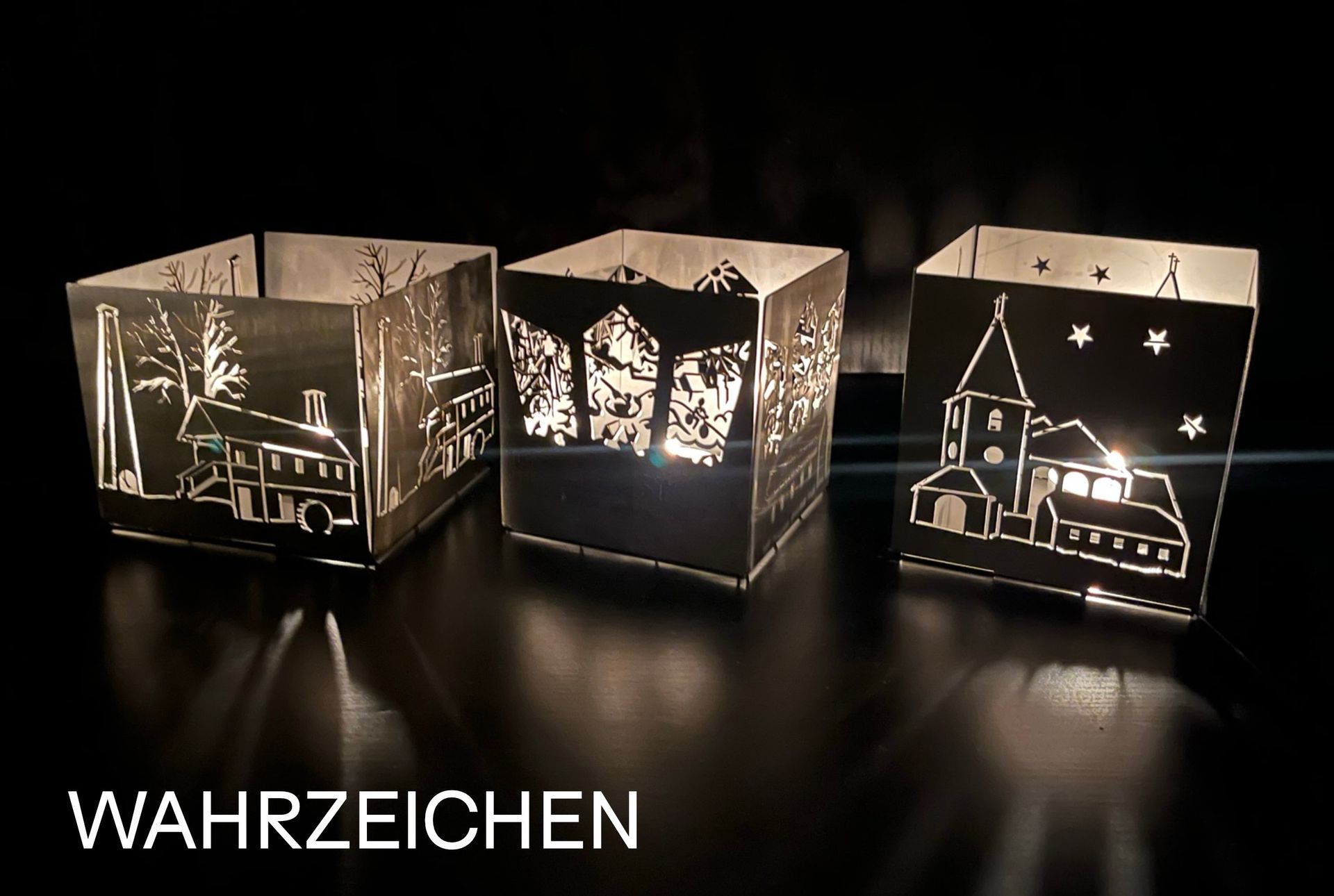 Drei beleuchtete, lasergeschnittene Metalllaternen mit Dorfmotiven auf dunklem Untergrund, Text: 