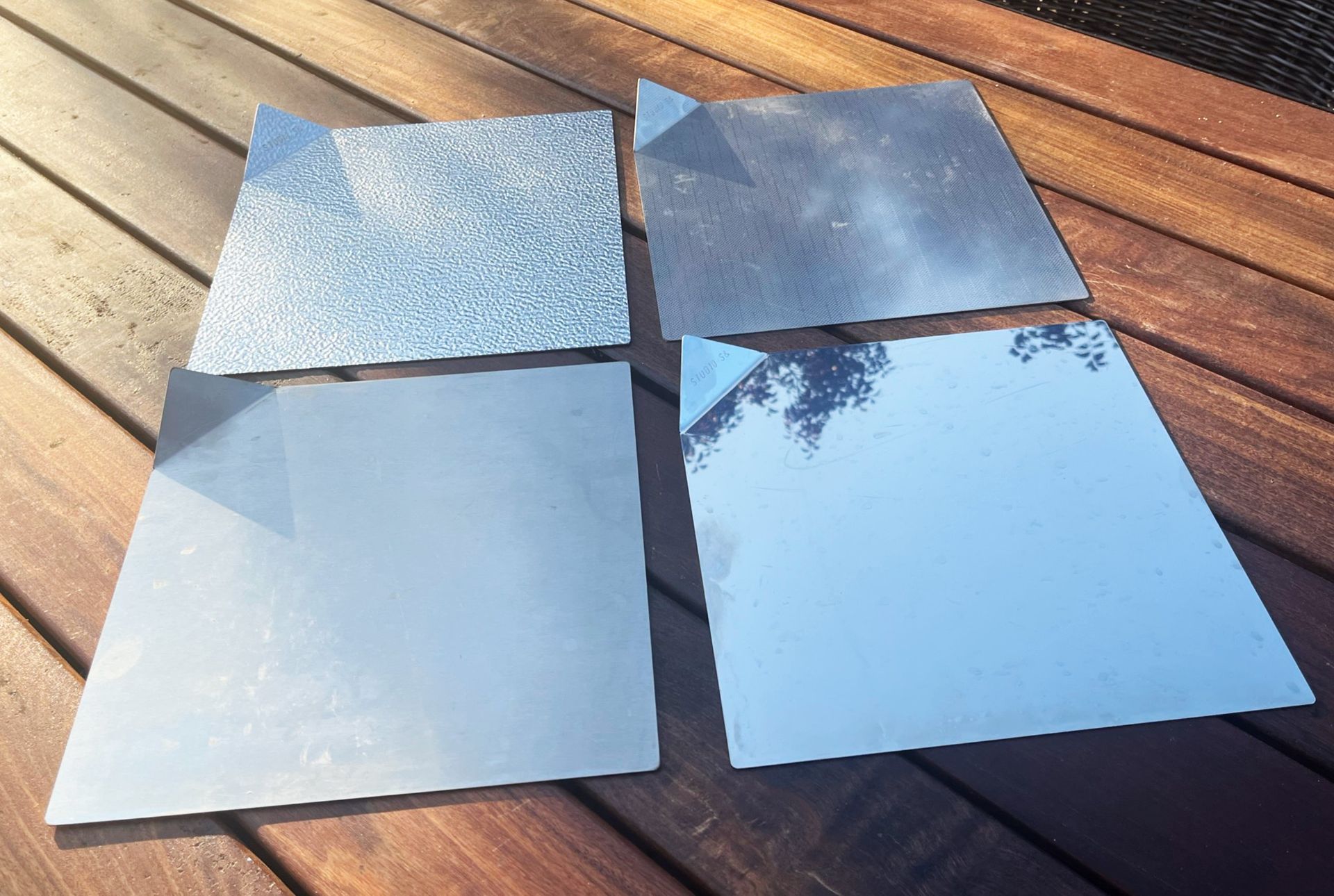 Vier quadratische Metallfliesen mit unterschiedlichen Oberflächenstrukturen, darunter eine mit Glitzer