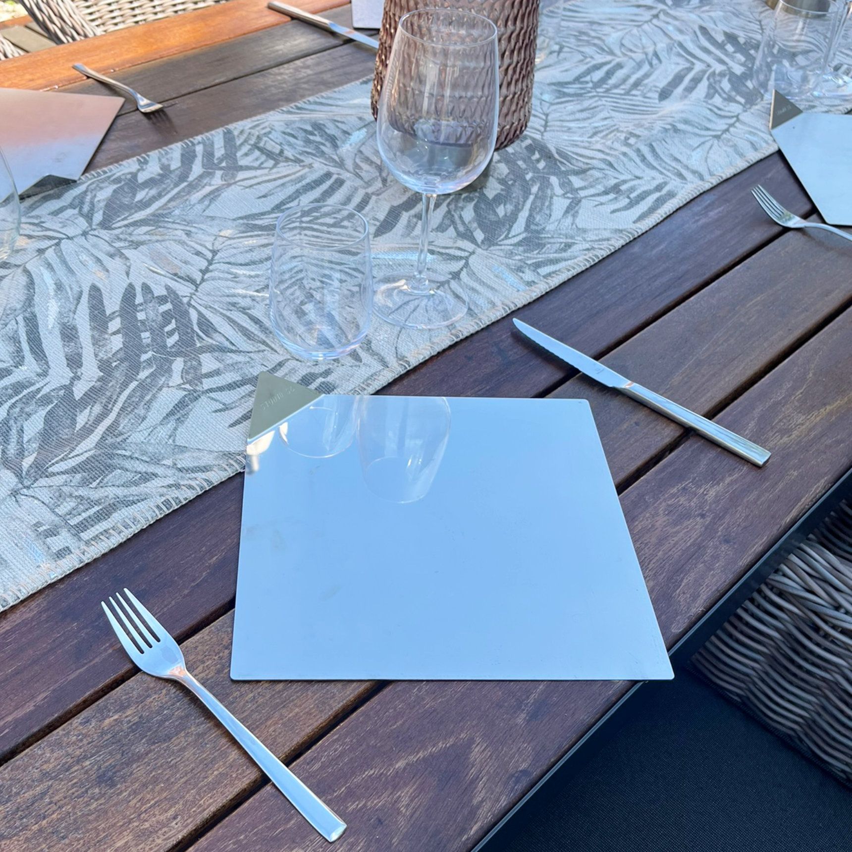 Tischdekoration mit einem spiegelähnlichen quadratischen Teller, Silberbesteck und Weinglas auf einem Holztisch