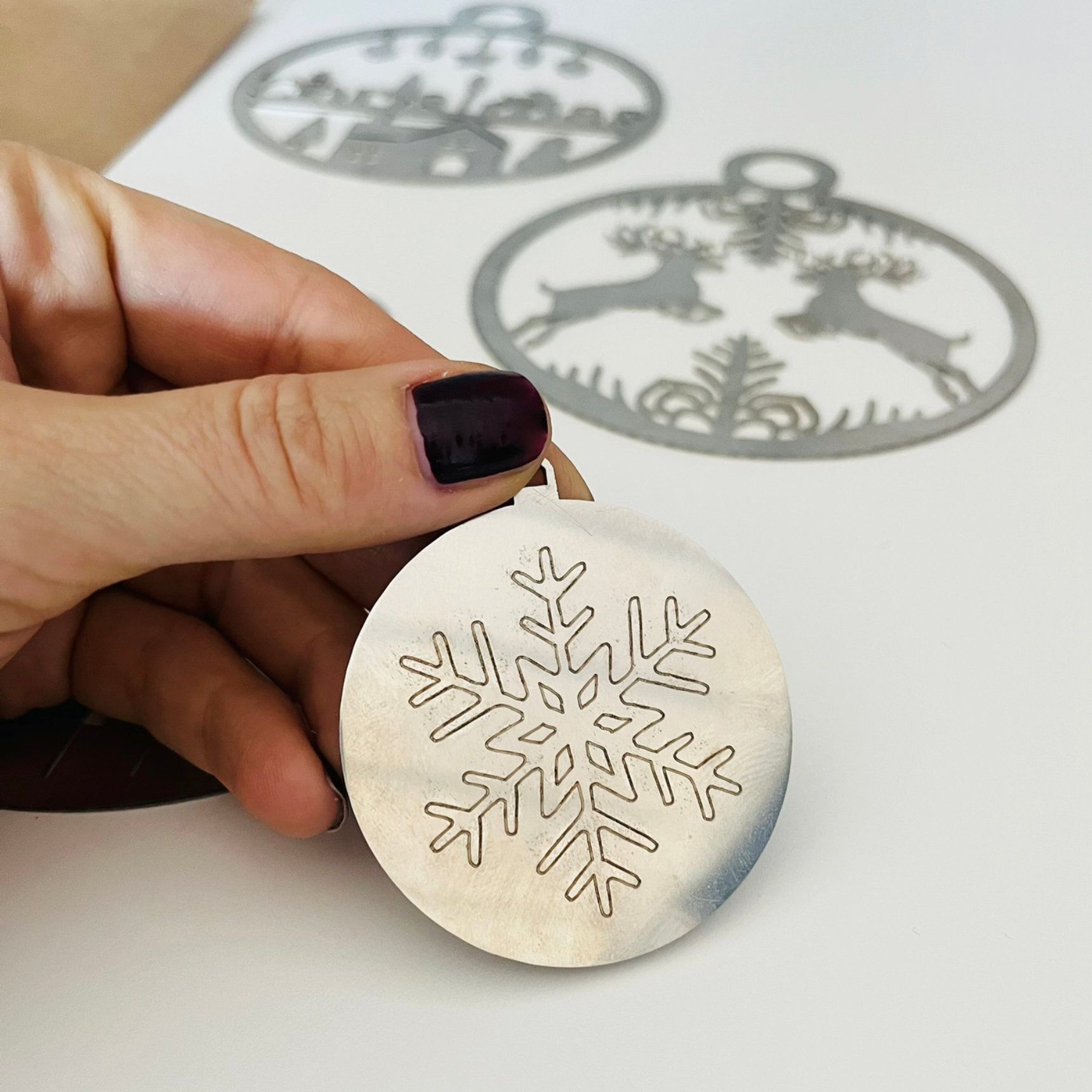 Eine Hand hält eine silberne Schneeflocke als Weihnachtsschmuck; im Hintergrund sind weitere Schmuckstücke zu sehen.