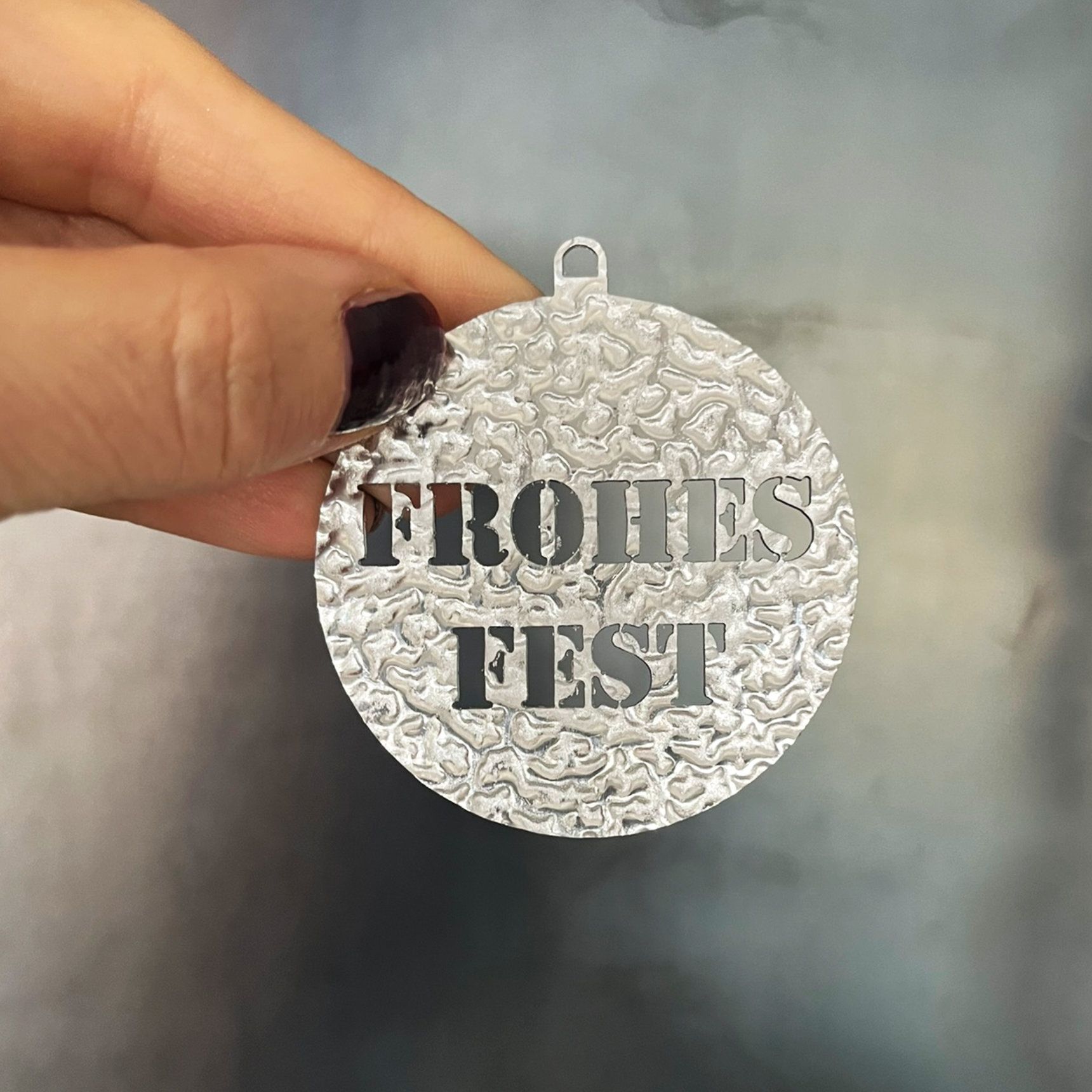 Eine Hand hält einen silbernen Schmuckanhänger mit der Aufschrift „FROHES FEST“.