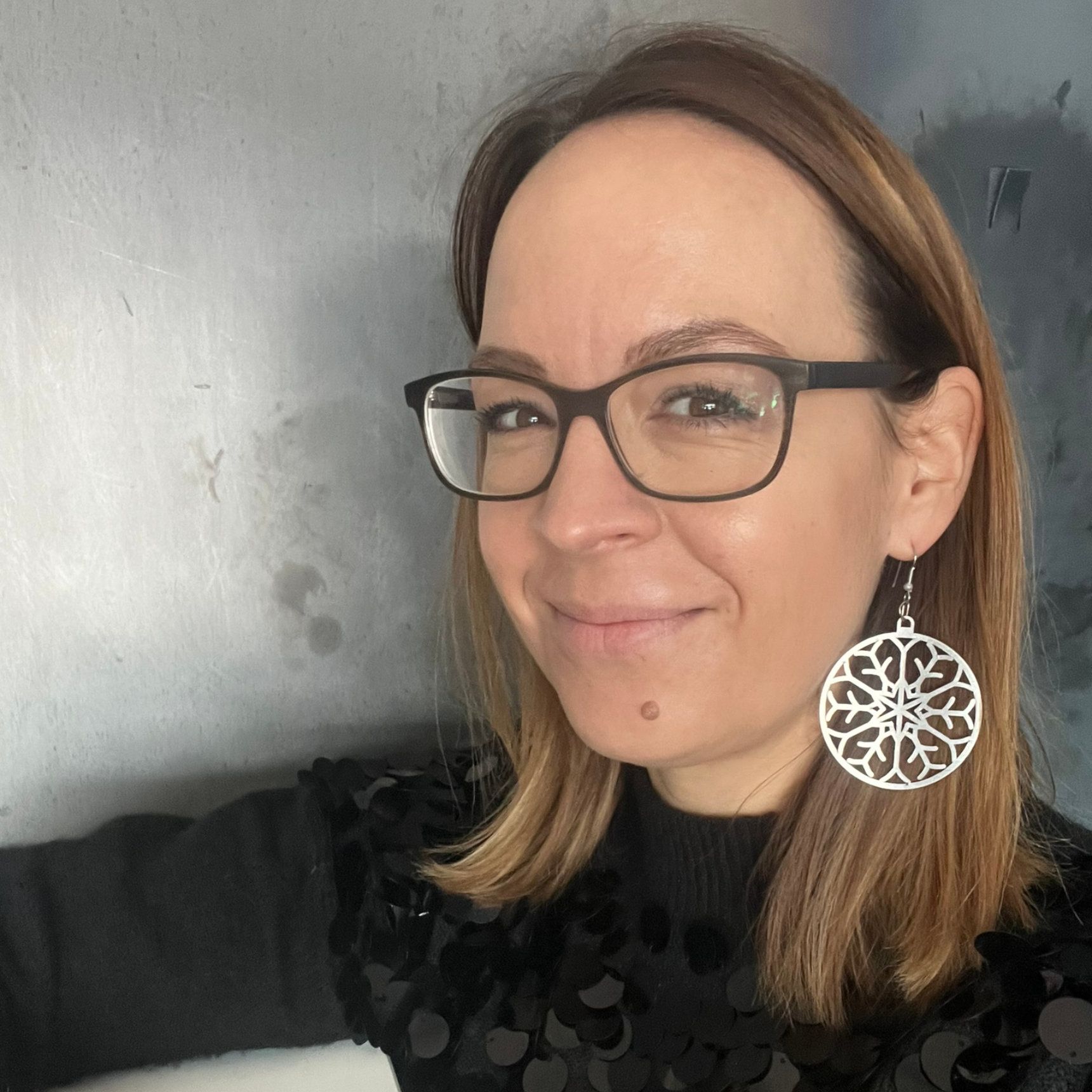 Frau mit Brille und großen silbernen Ohrringen, lächelnd. Lehnt sich an eine silberne Wand.