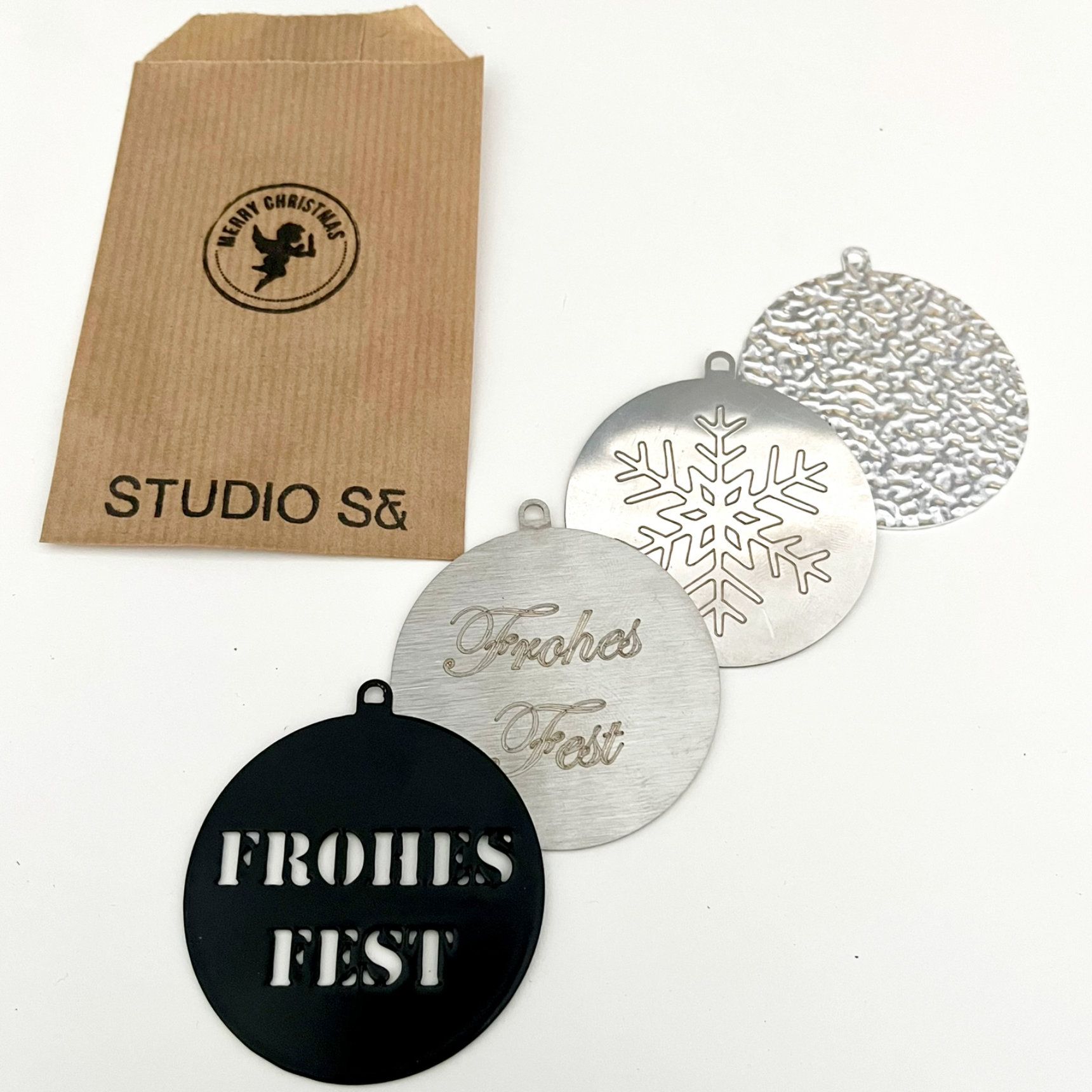 Vier silberne Weihnachtskugeln und eine Papiertüte. Auf einer Kugel steht „Frohes Fest“, eine andere ist eine Schneeflocke.