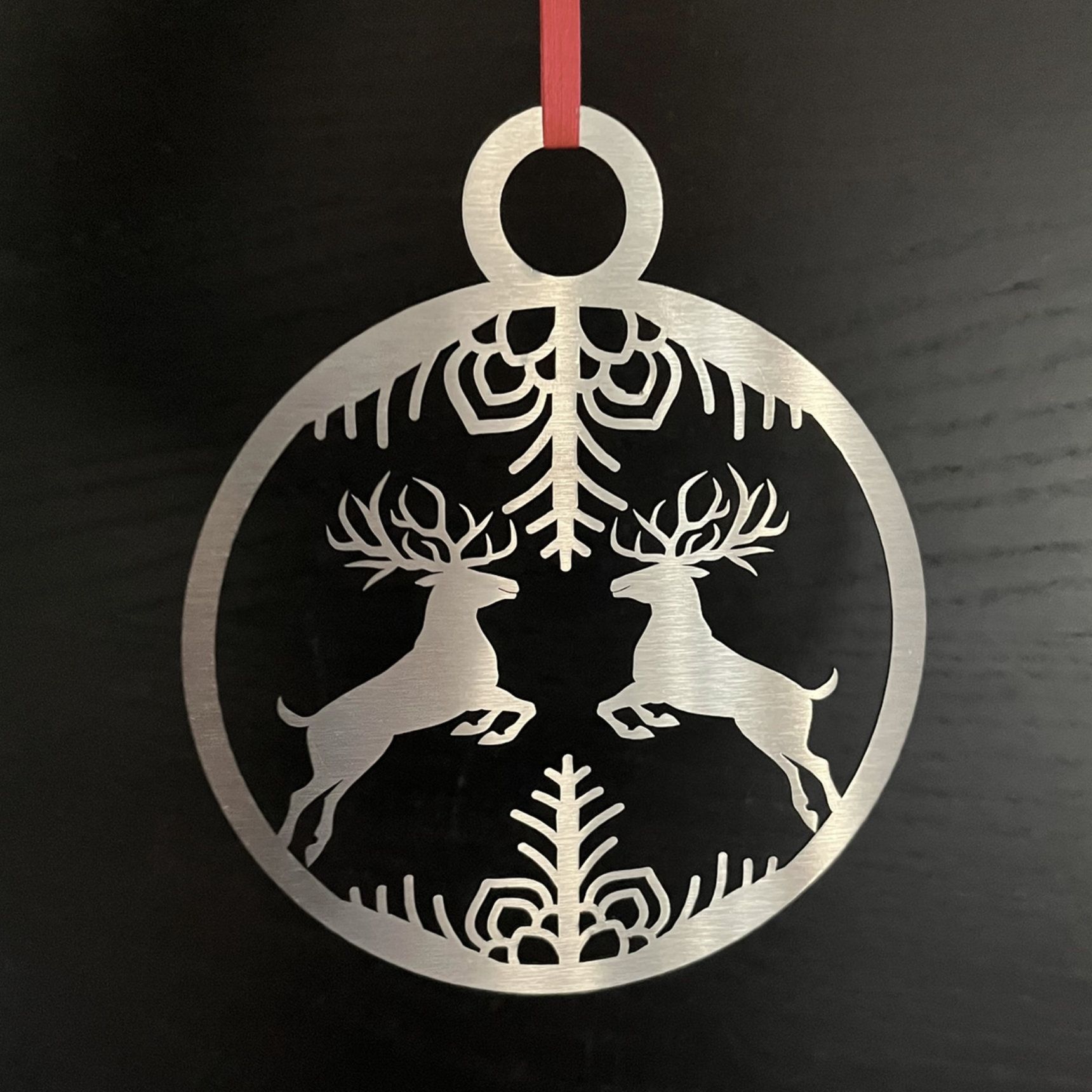 Silberner Weihnachtsschmuck mit zwei springenden Hirschen und Schneeflockenmotiv, rotes Band.