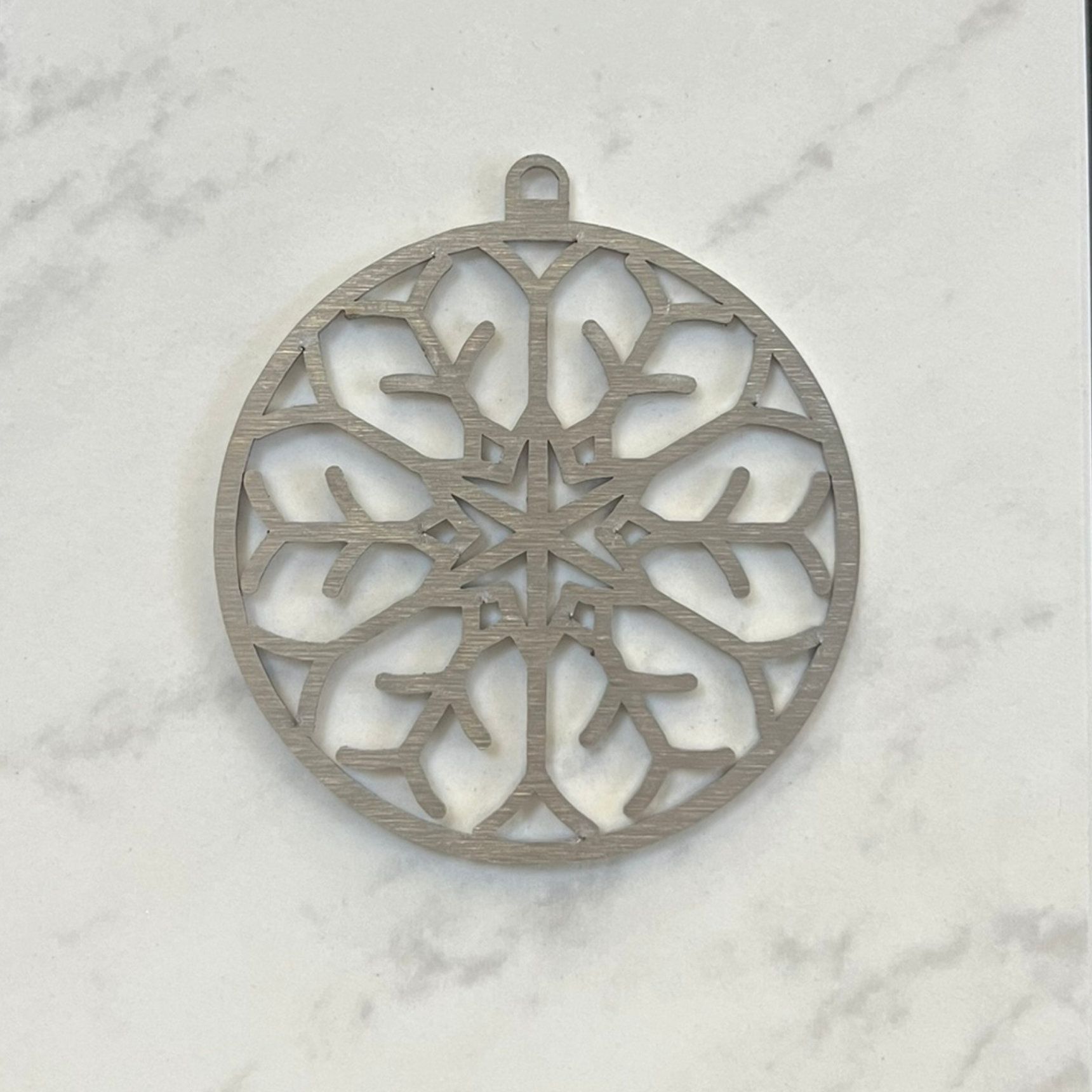 Silberne, lasergeschnittene Schneeflocken-Dekoration aus Holz. Rund mit Aufhängeöse. Auf Marmorgrund.