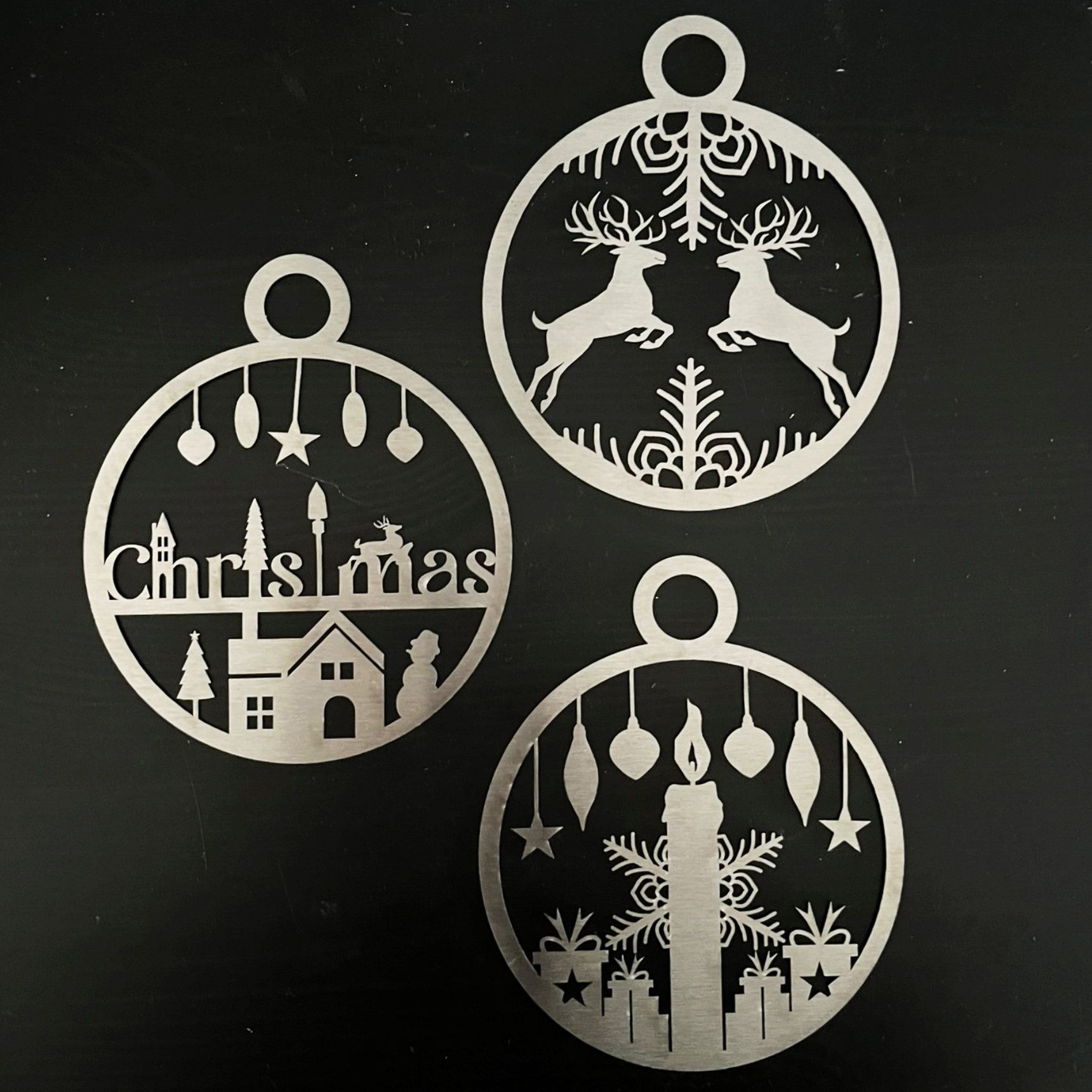 Drei silberne Weihnachtsornamente mit ausgeschnittenen Weihnachtsmotiven, jeweils mit einer Schlaufe zum Aufhängen.