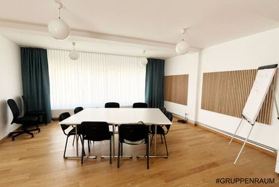 Besprechungsraum mit weißem Tisch, schwarzen Stühlen, Holzboden, türkisfarbenen Vorhängen und einer Whiteboard-Tafel.