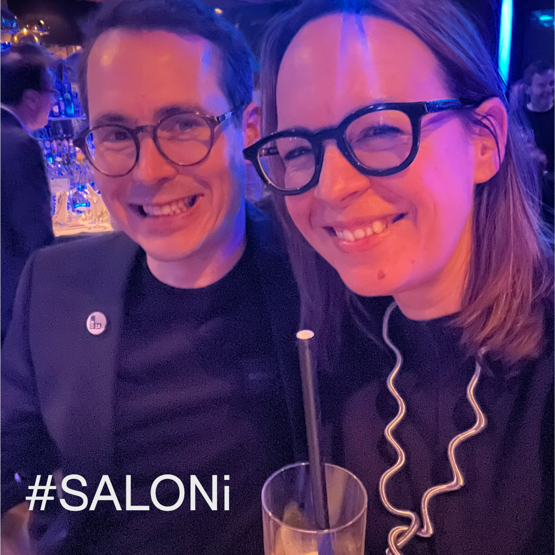 Zwei lächelnde Personen mit Brille; eine davon trägt einen schwarzen Blazer, ein Getränk und ein "#SALONI"-Schild.