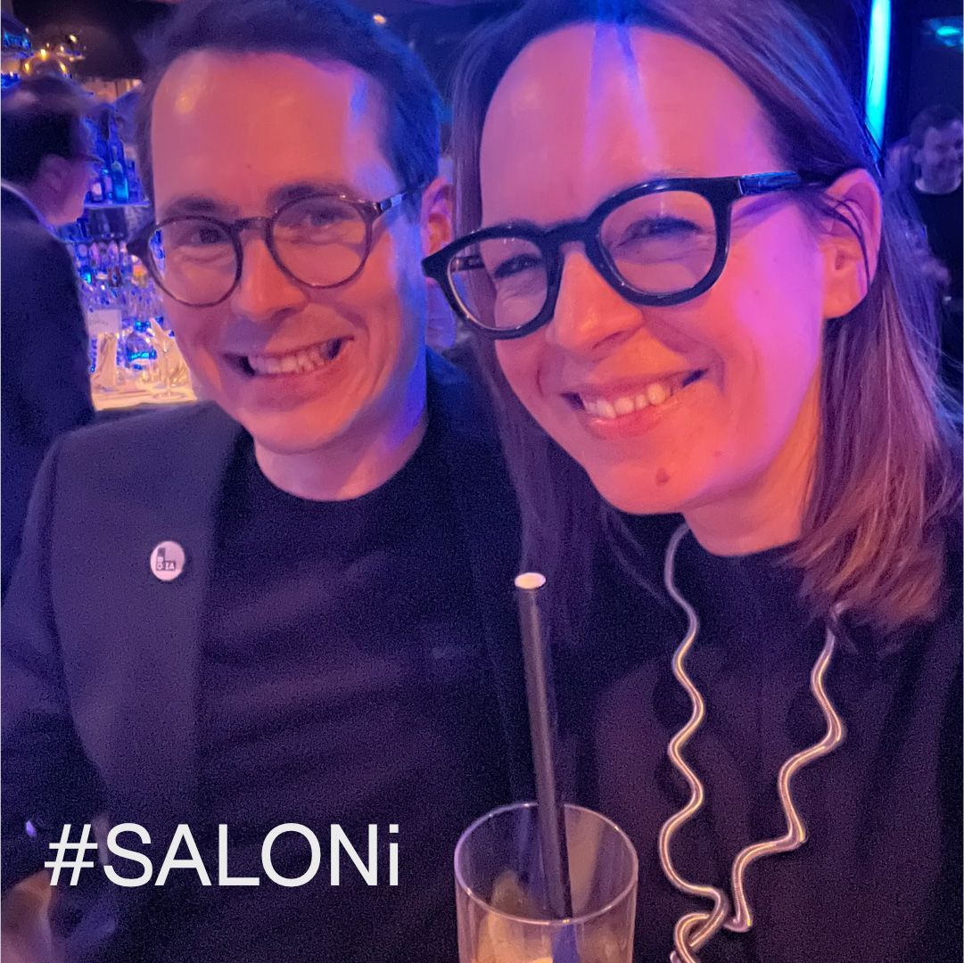 Zwei lächelnde Personen mit Brille; eine davon trägt einen schwarzen Blazer, ein Getränk und ein "#SALONI"-Schild.