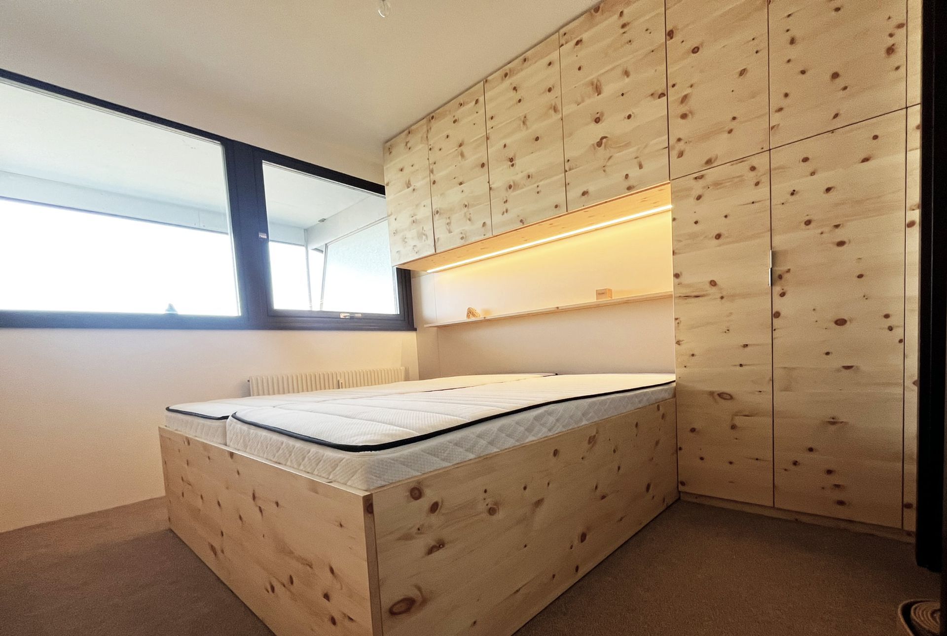 Schlafzimmer mit Holzbett, Kopfteil und Einbauschrank. Großes Fenster links.