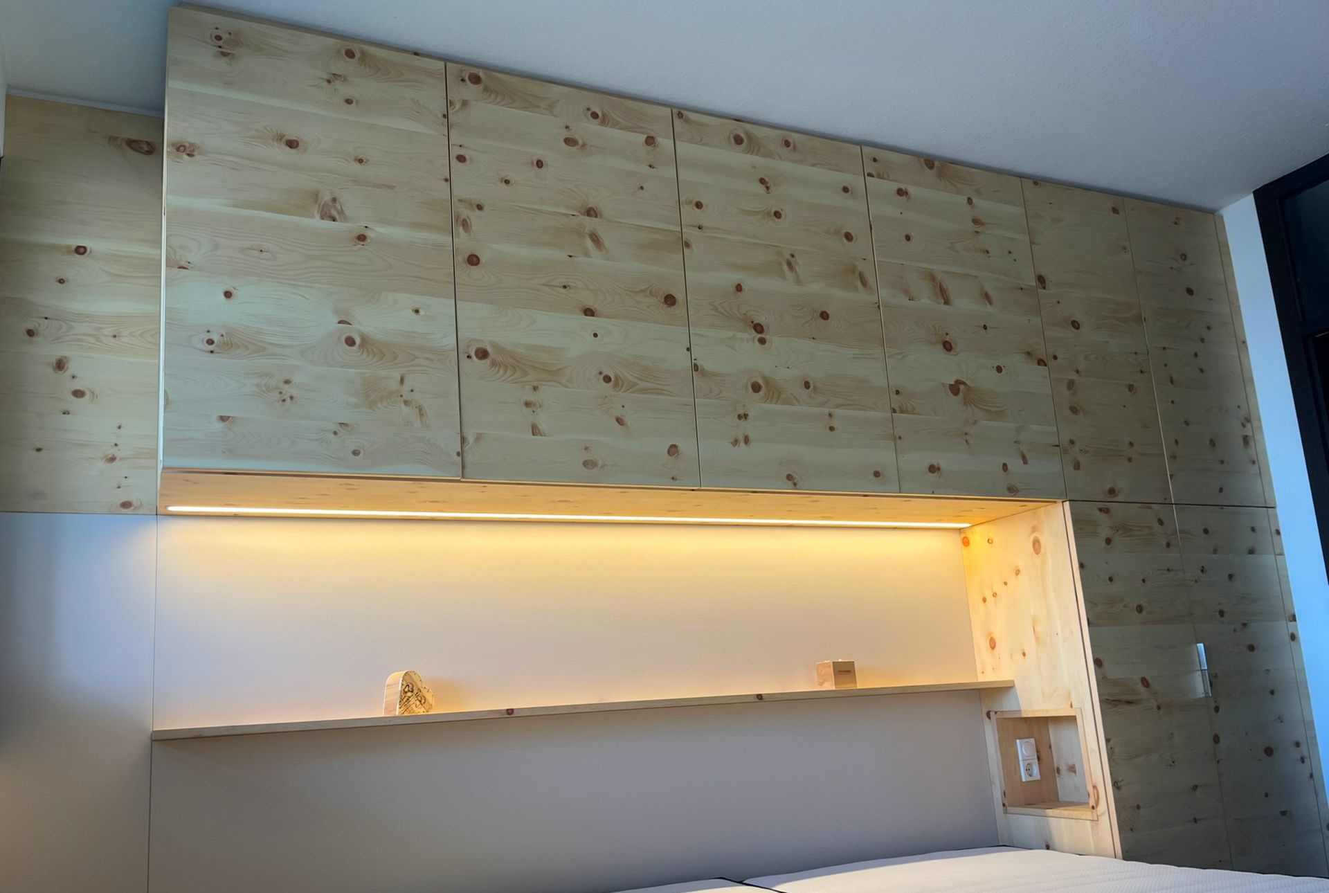 Holzwand mit integriertem Bettkopfteil und Regalen, beleuchtet mit warmem Licht.