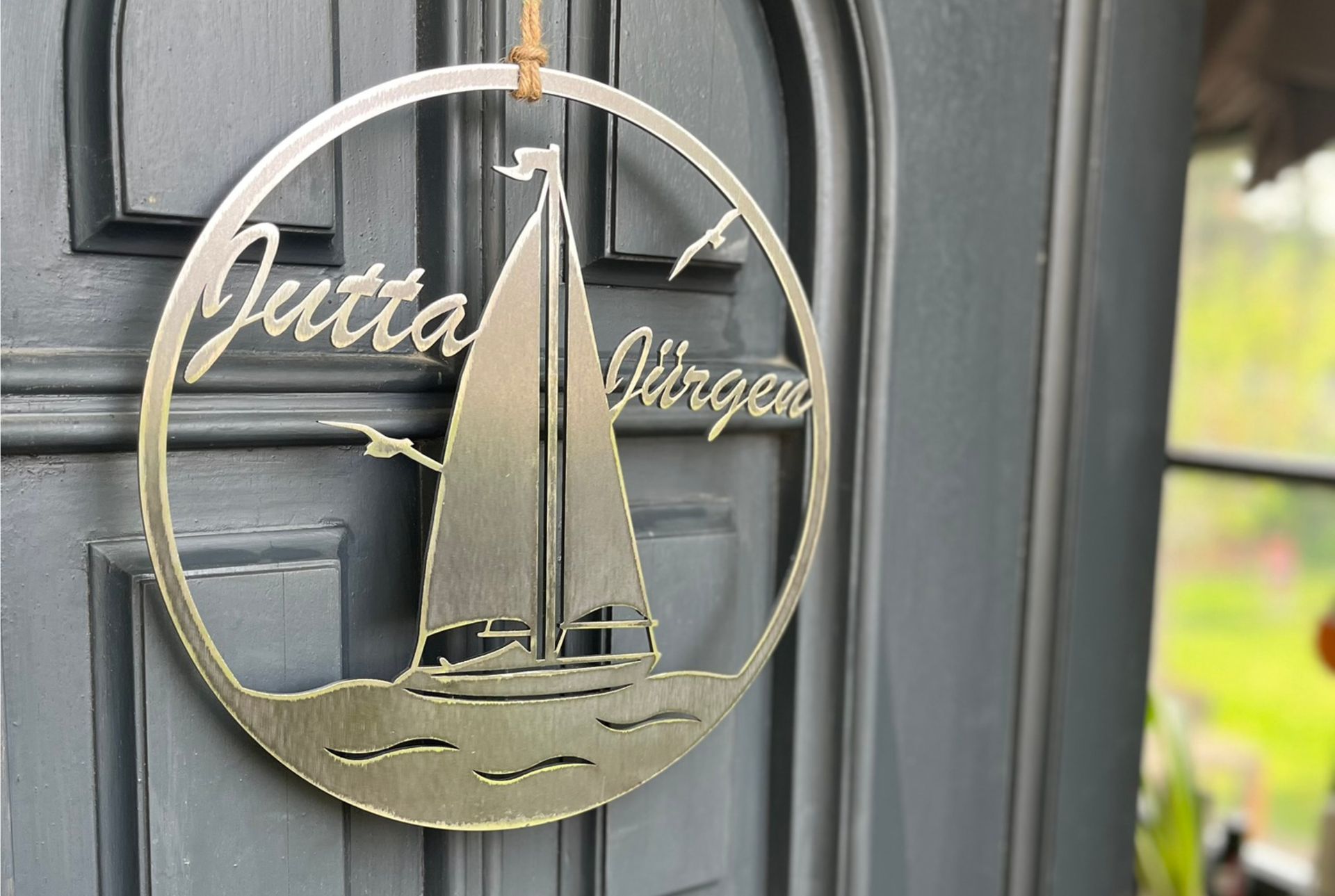 Ein rundes Metallschild an einer blauen Tür mit einem Segelboot und den Namen „Gutta“ und „Jürgen“.