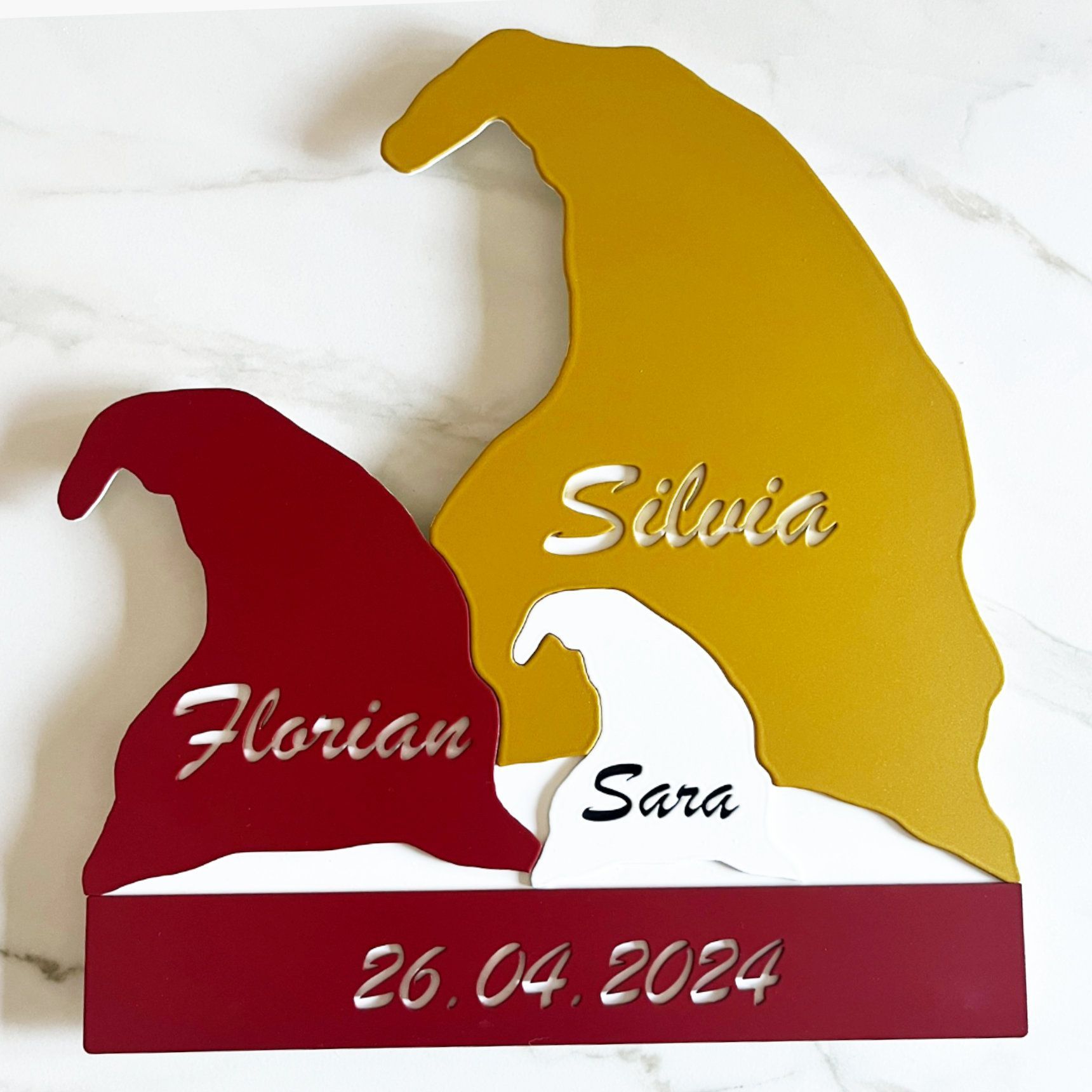 Familienschild mit Zwergenmotiven. Rote, goldene und weiße Zwerge mit den Namen „Florian“, „Silvia“, „Sara“ und dem Datum.