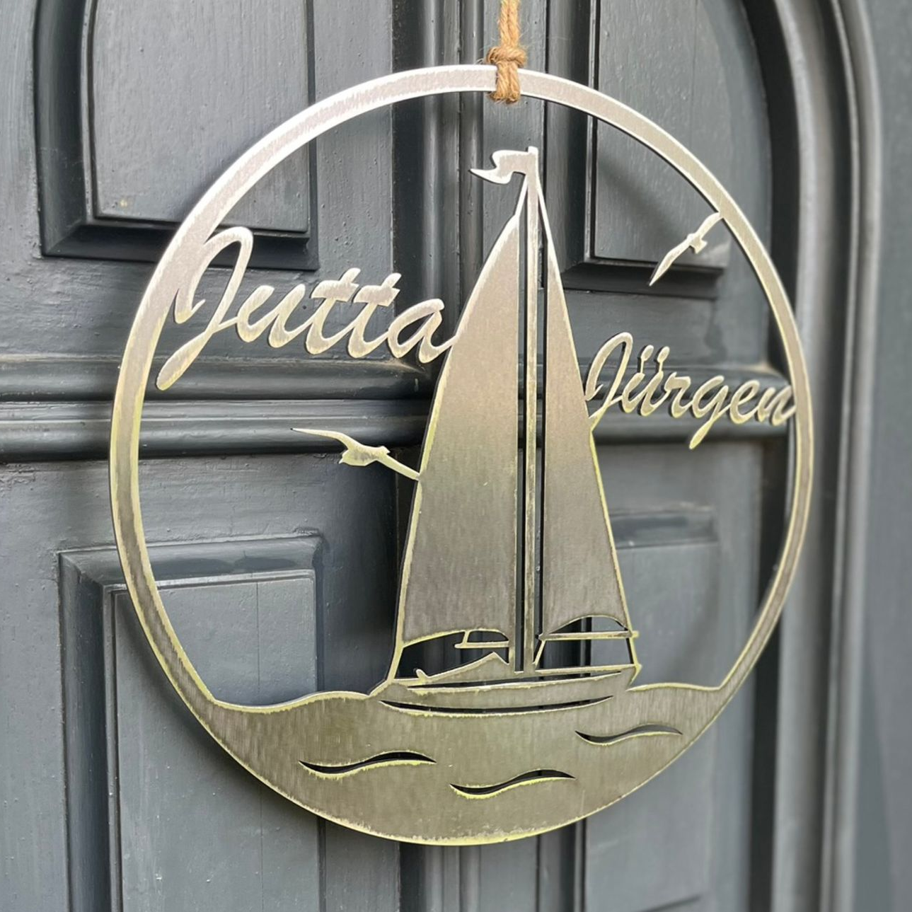 Ein rundes Metallschild mit den Namen „Jutta“ und „Jürgen“, einem Segelboot und einer Möwe hängt an einer grauen Tür.