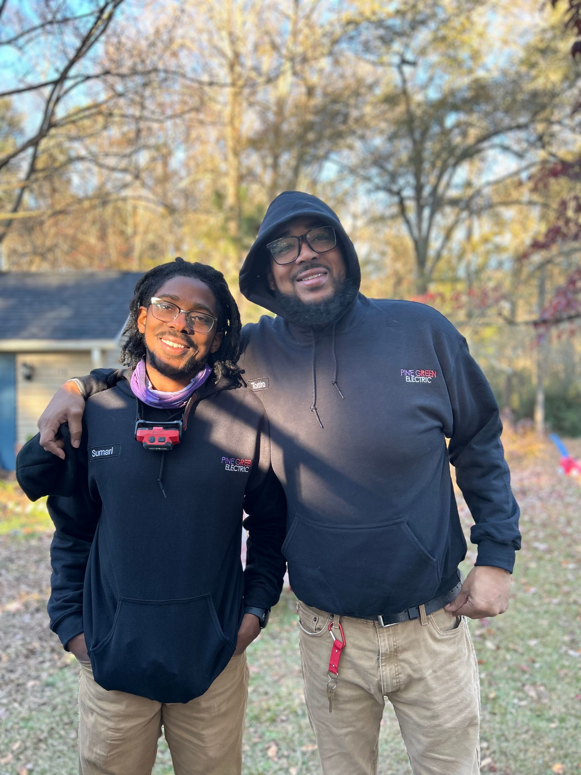 Two people outdoors, one with arm around the other. Both wear black hoodies and tan pants. Smiling, outdoor setting.