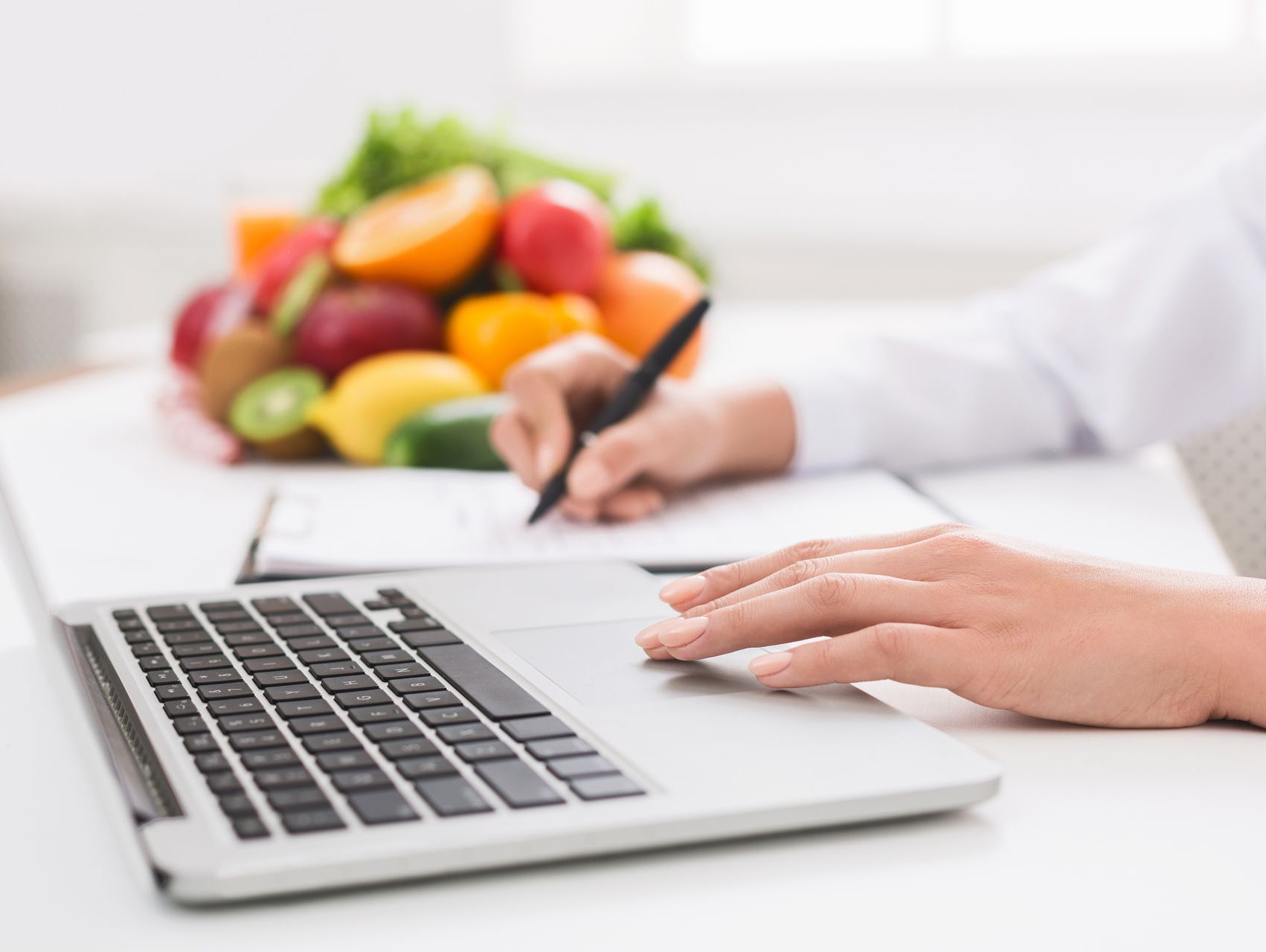 A Doctor in a White Coat Typing at a Desk with a Fruit Bowl