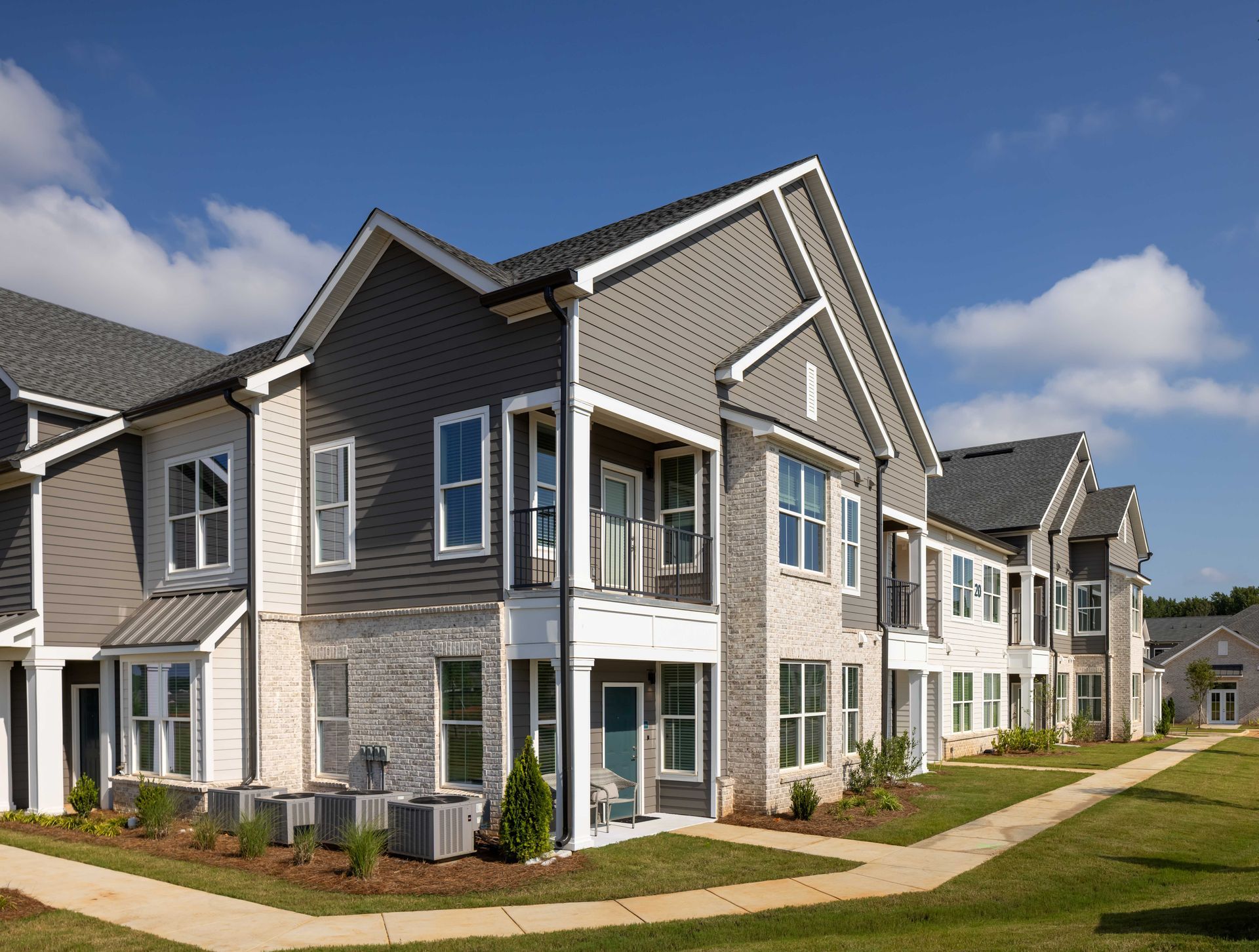 Two-story apartment building with gray siding, brick, and white trim on a sunny day.