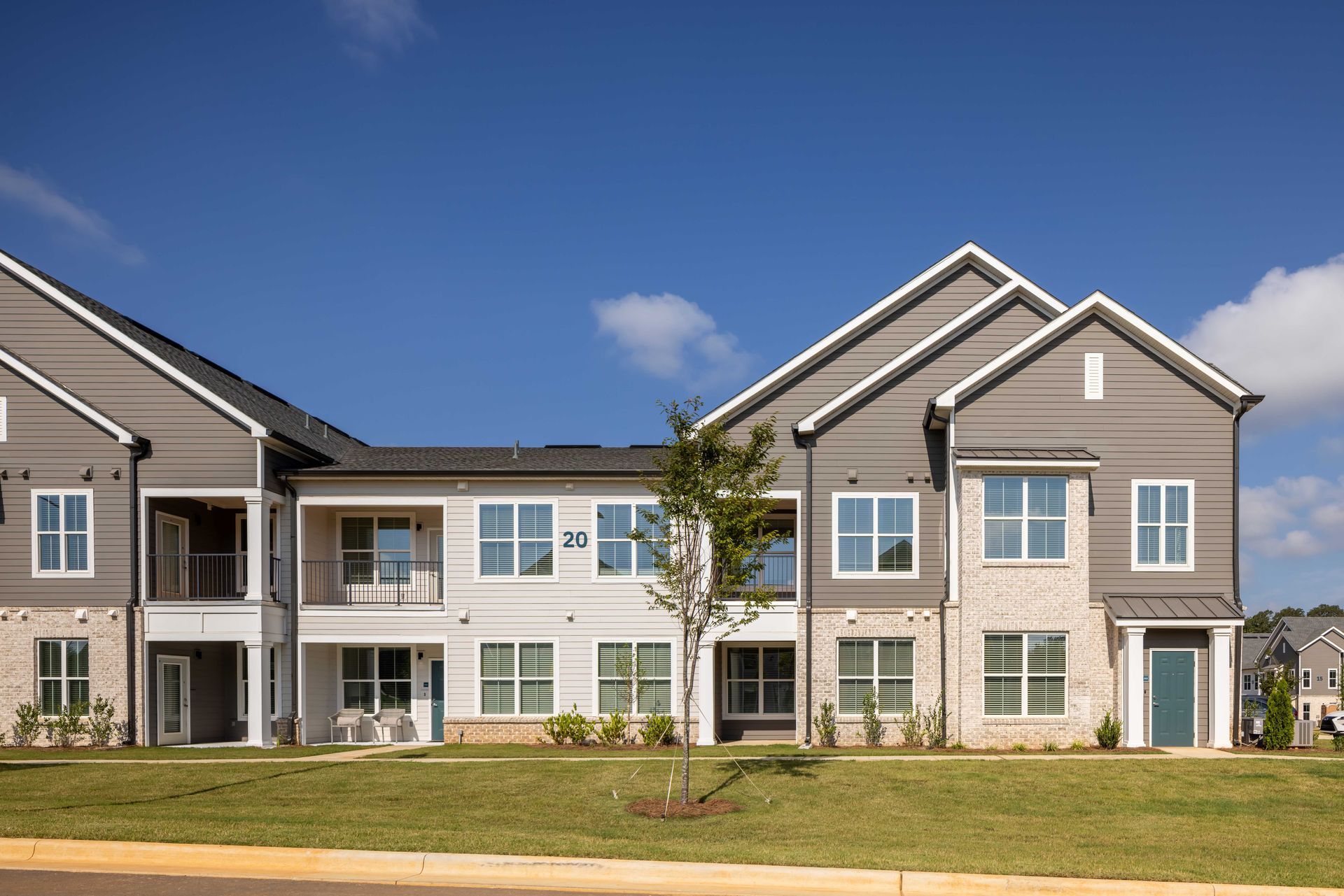 Multi-unit building with tan brick, grey siding, blue door, and green lawn under a blue sky.