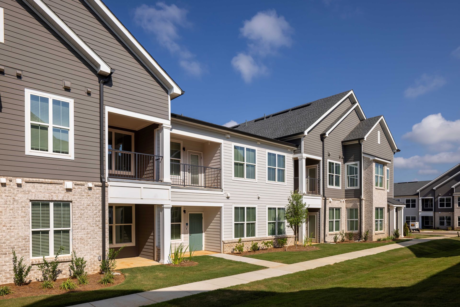 Exterior view of a two-story apartment complex with gray siding, brick accents, and a sunny sky.