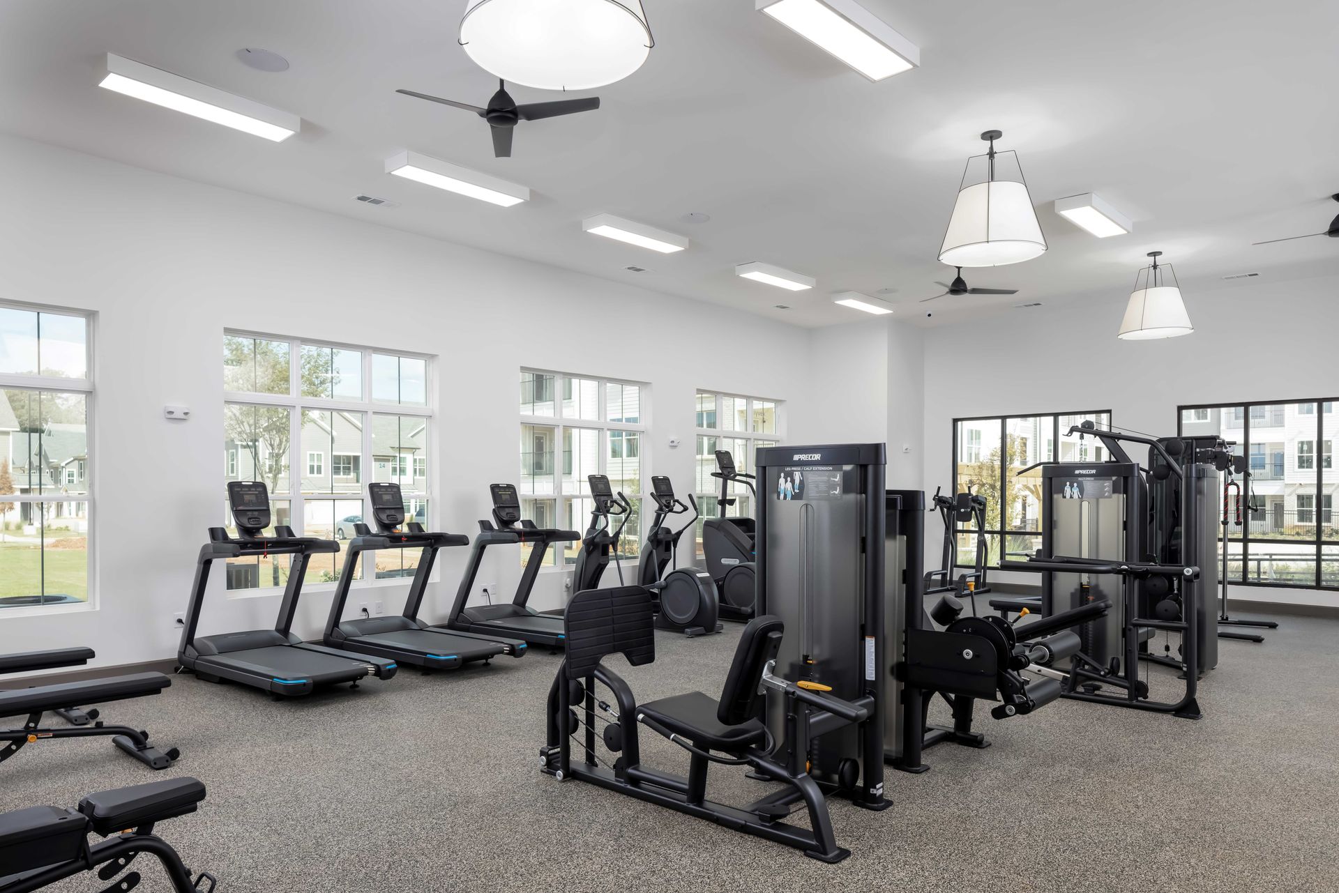 A modern gym with treadmills, weight machines, and exercise equipment, lit by white lights and windows.