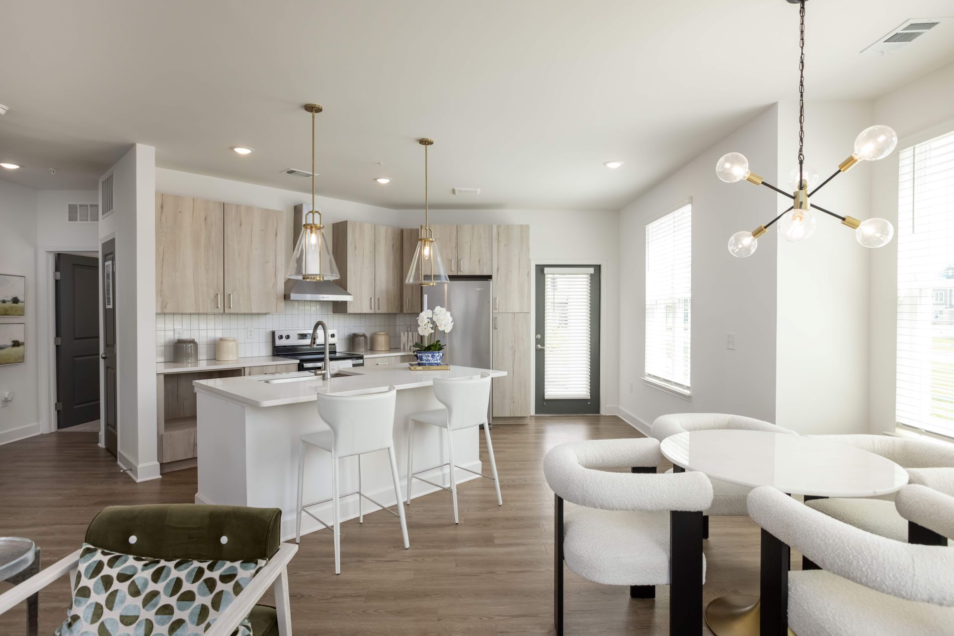 Modern apartment lounge with seating, a kitchen area, and large chandelier.