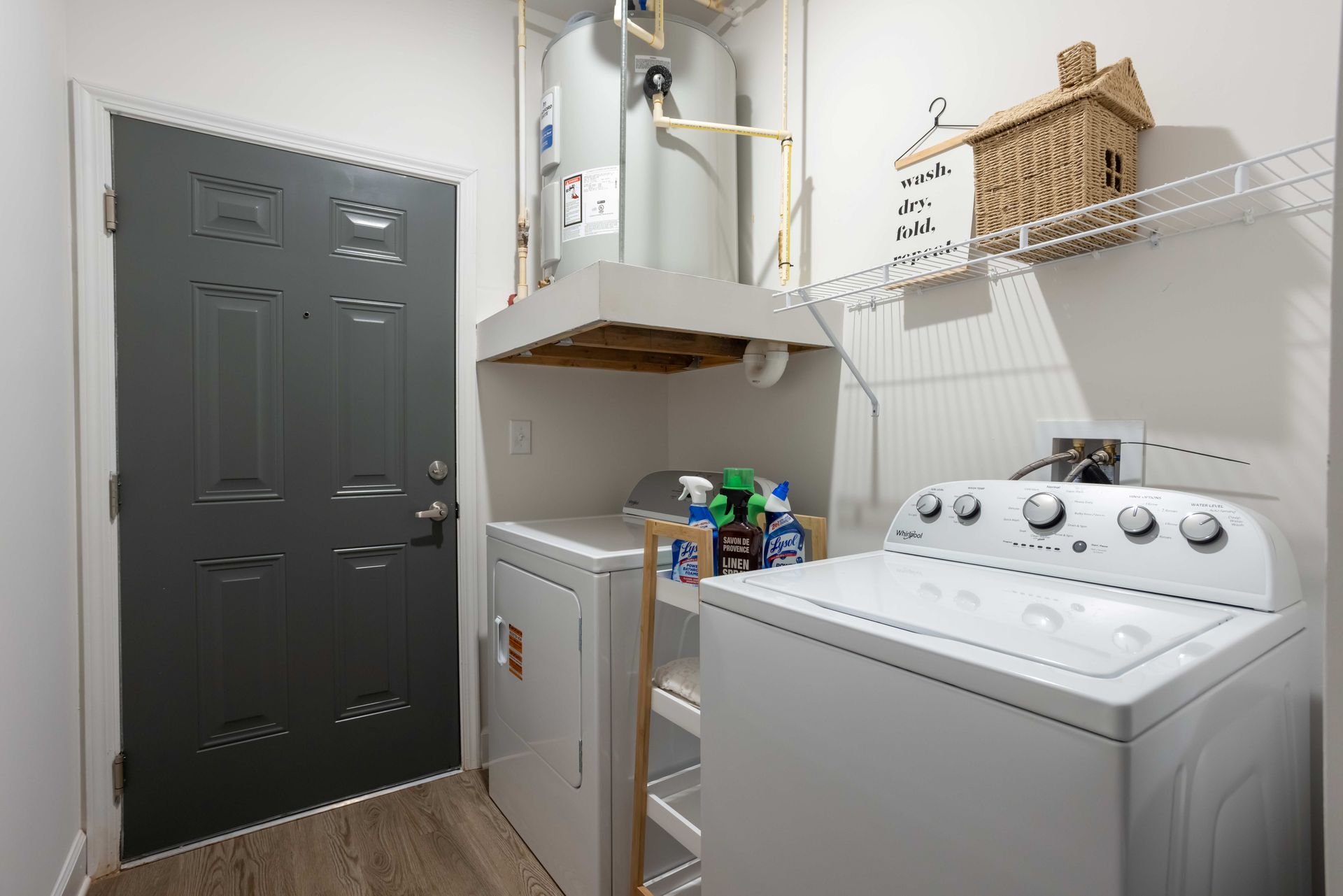 Laundry room with a white washer, dryer, and water heater. Gray door, wooden shelf, and cleaning supplies.