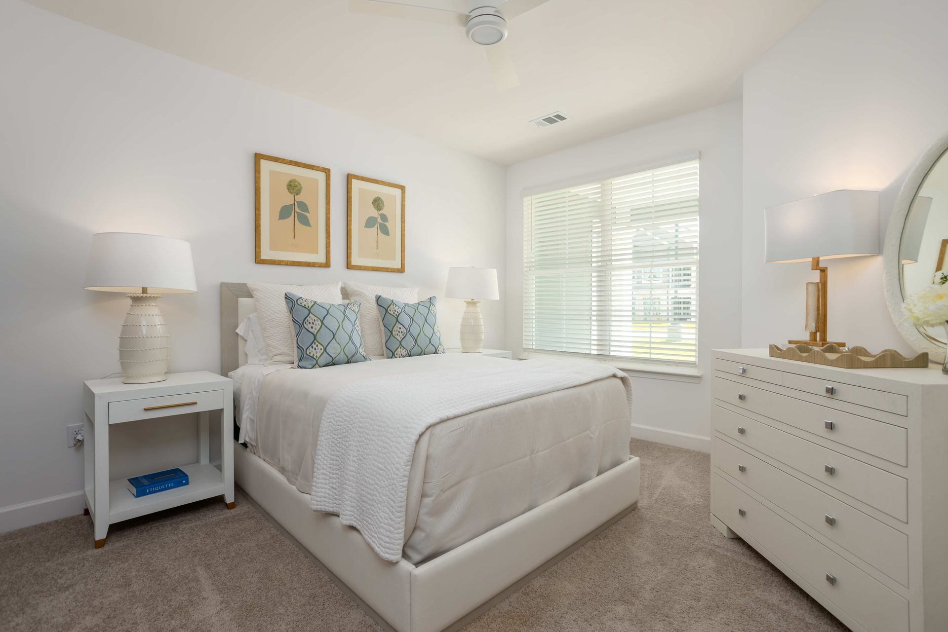 Bright bedroom with white walls, bed, and dresser, floral artwork, and a window with blinds.