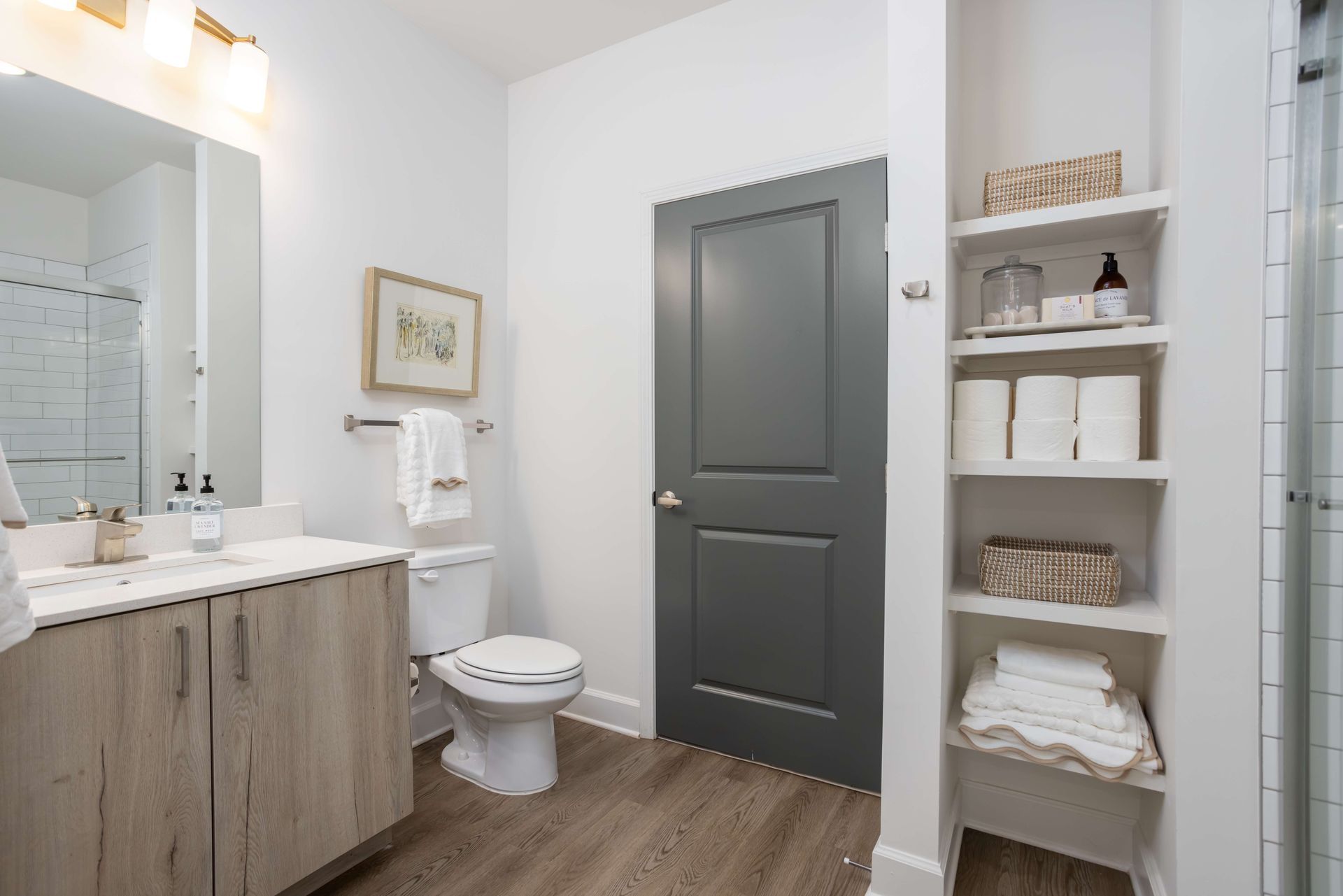 Bathroom with wooden floor, white walls, and grey door; includes vanity, toilet, and shelves with linens.