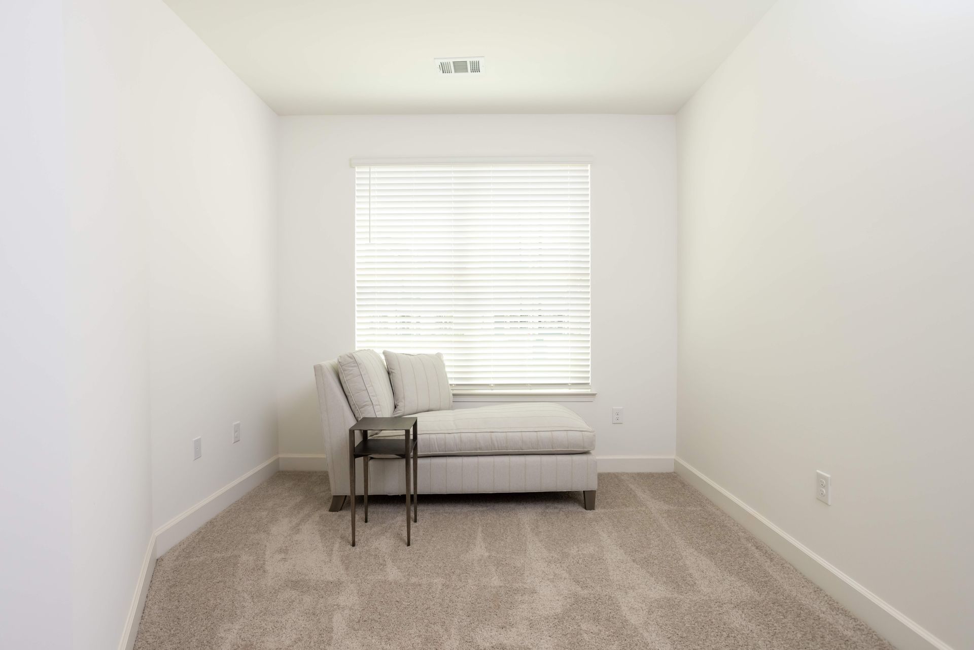 A small, empty room with a chaise lounge and a side table in front of a window.