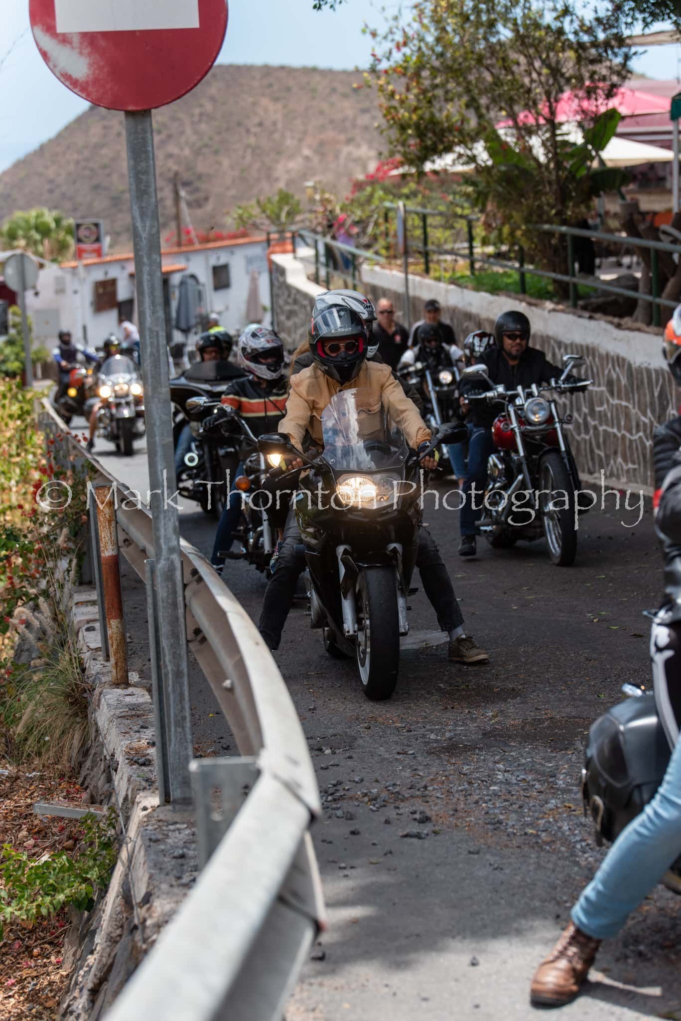 A group of people are riding motorcycles down a road with a no entry sign in the background