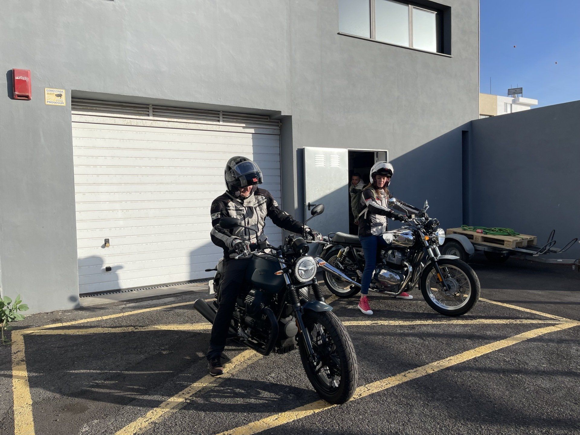 Two men are riding motorcycles in a parking lot in front of a building.